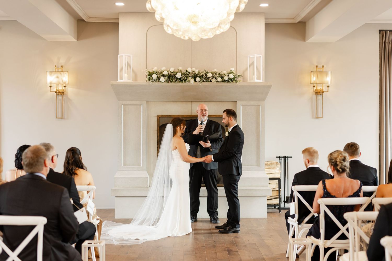 Bride and groom exchange vows at elegant indoor ceremony with floral fireplace mantel and chandelier.