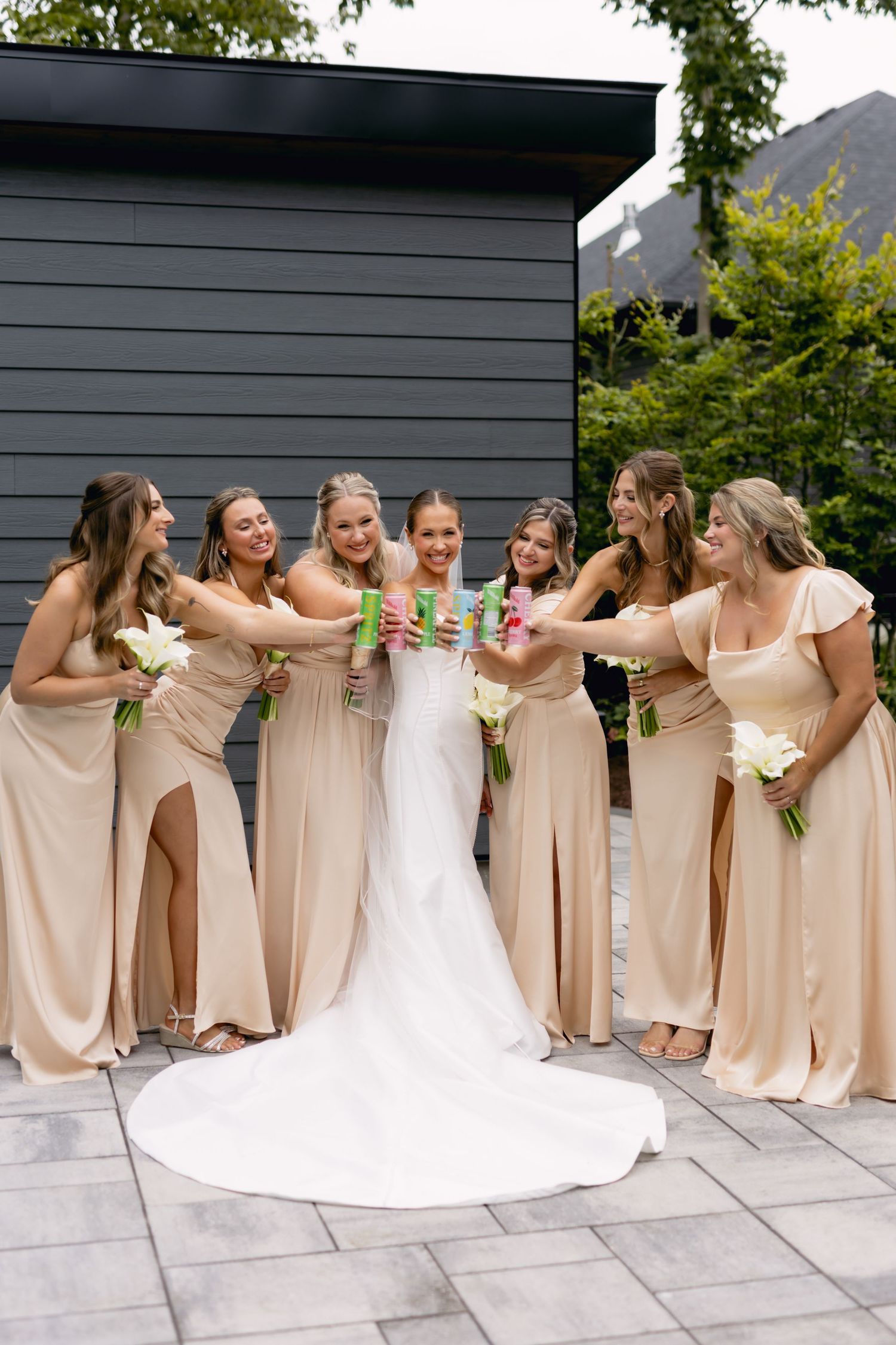 Bride in white gown toasts with six bridesmaids in champagne dresses holding white floral bouquets outdoors.