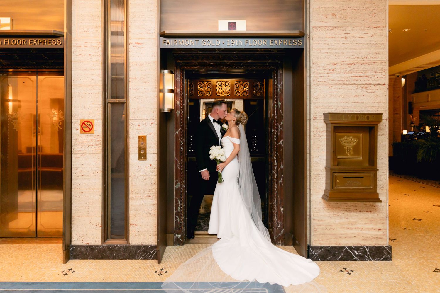 Wedding couple kissing in ornate Art Deco elevator lobby, bride in sleek white gown with long train.