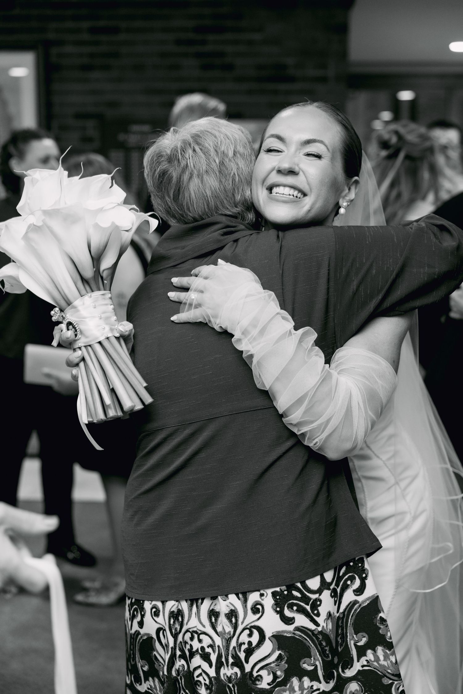 Black and white photo of a smiling bride hugging an elderly woman while holding a bouquet at a wedding reception.