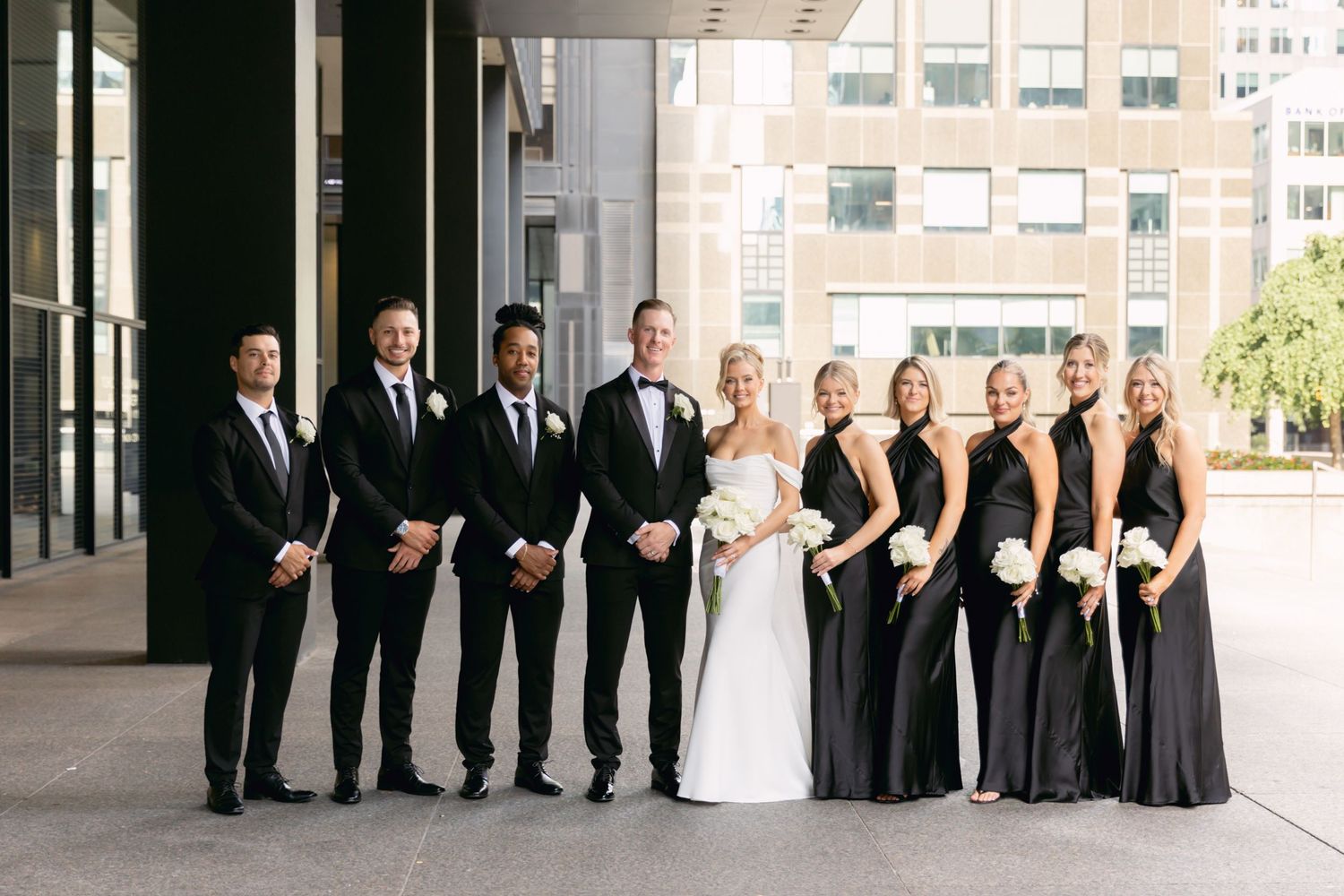 Wedding party posing in black attire outside modern building, bridesmaids holding white bouquets.
