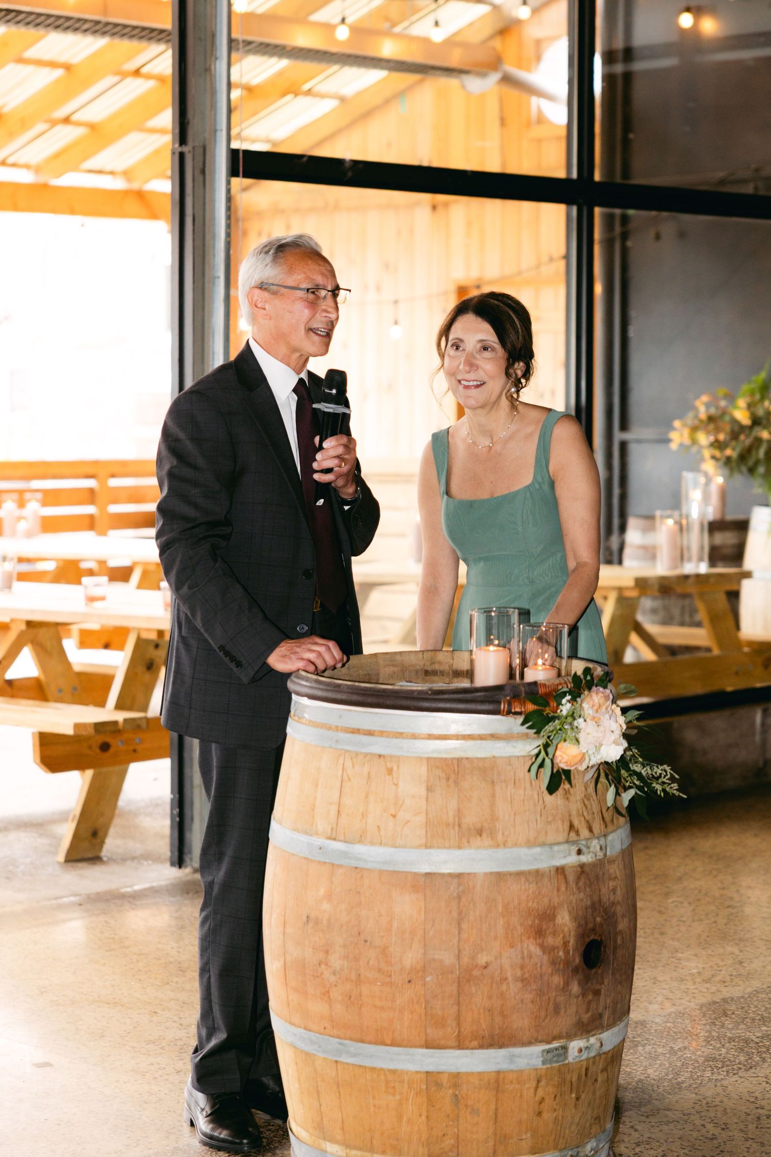Wedding guests gather around wooden barrels in a rustic venue with natural lighting.