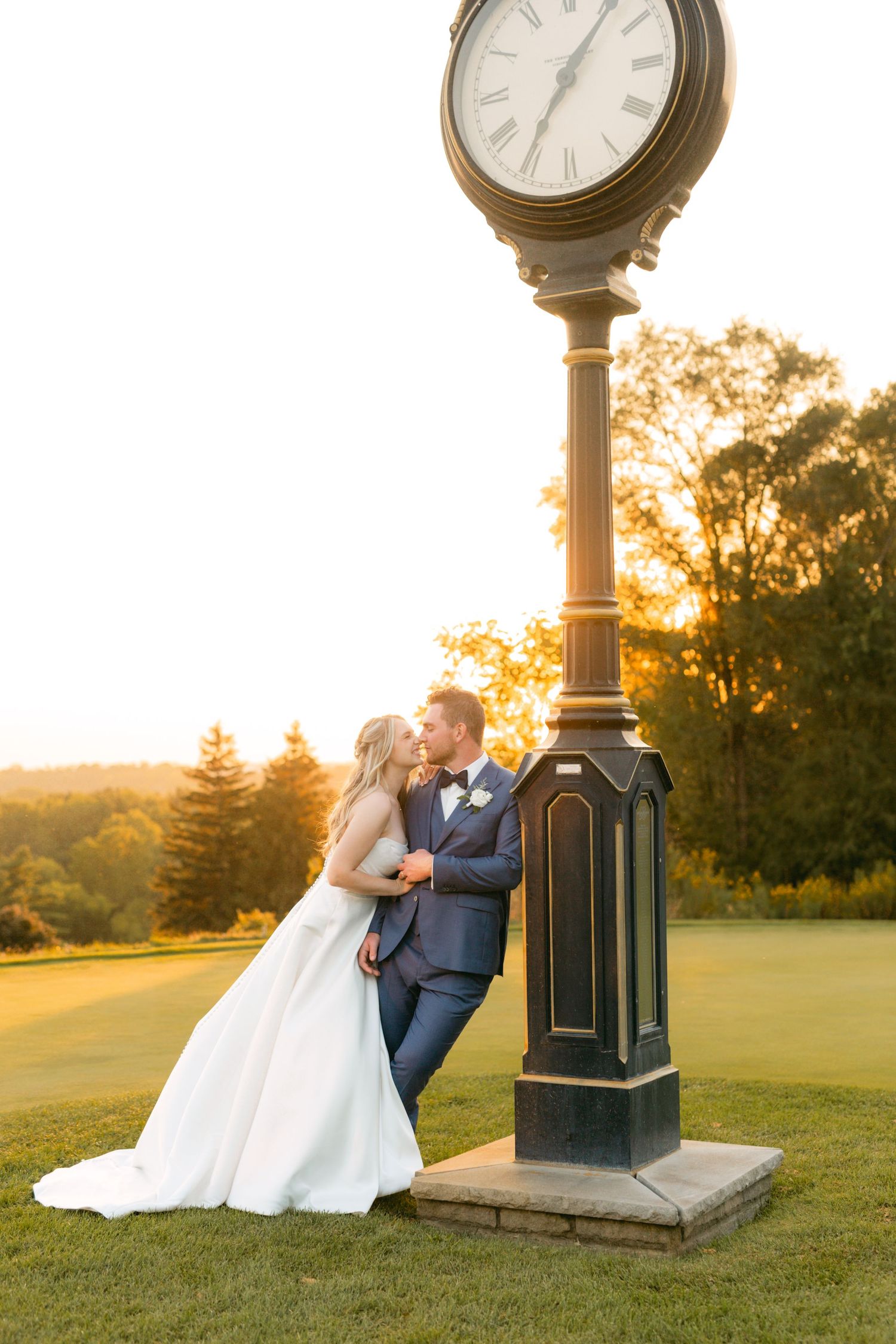 Wedding couple shares intimate moment near decorative clock post during golden hour on grassy hillside.