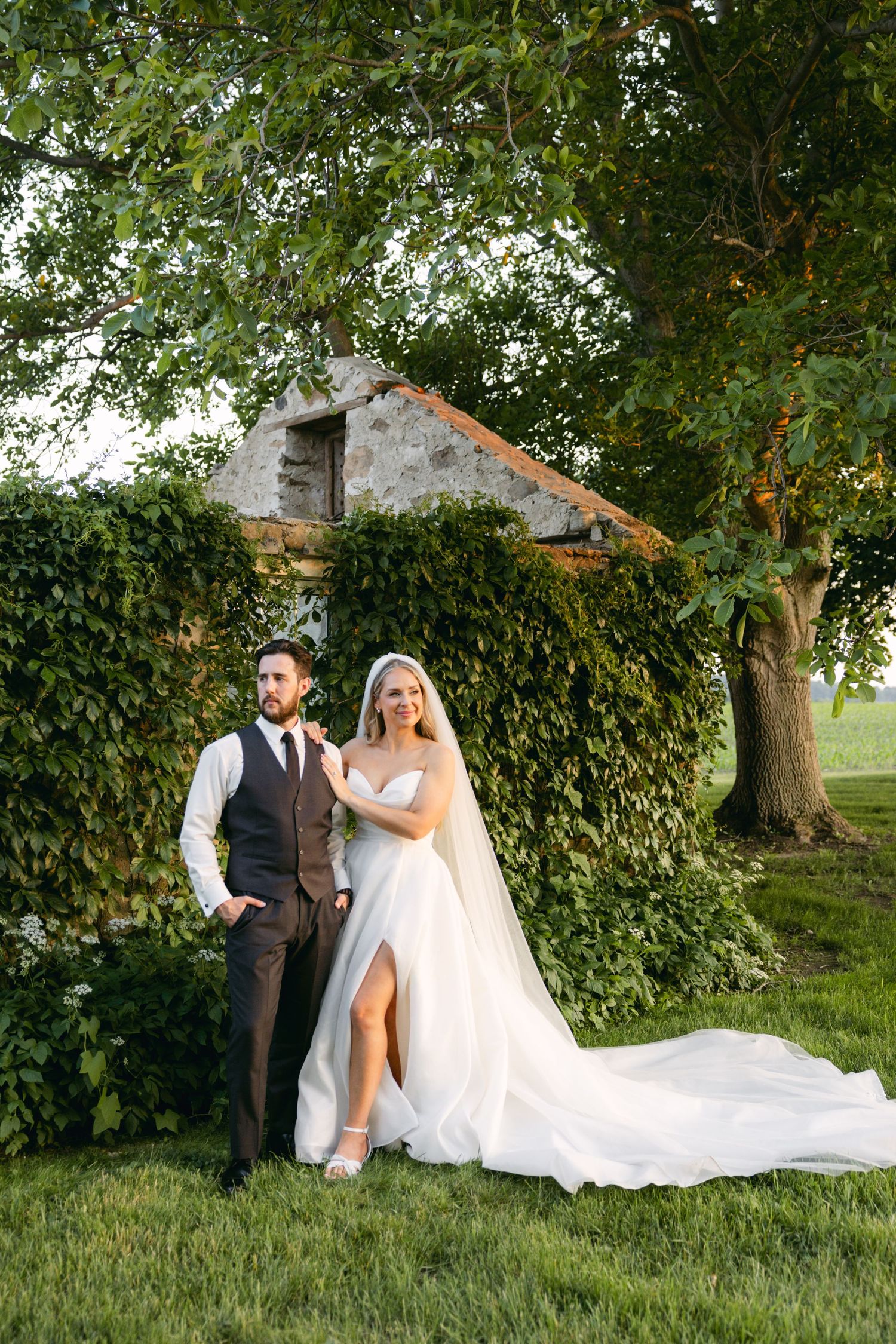 Wedding couple poses by ivy-covered wall as dress train flows in evening breeze.