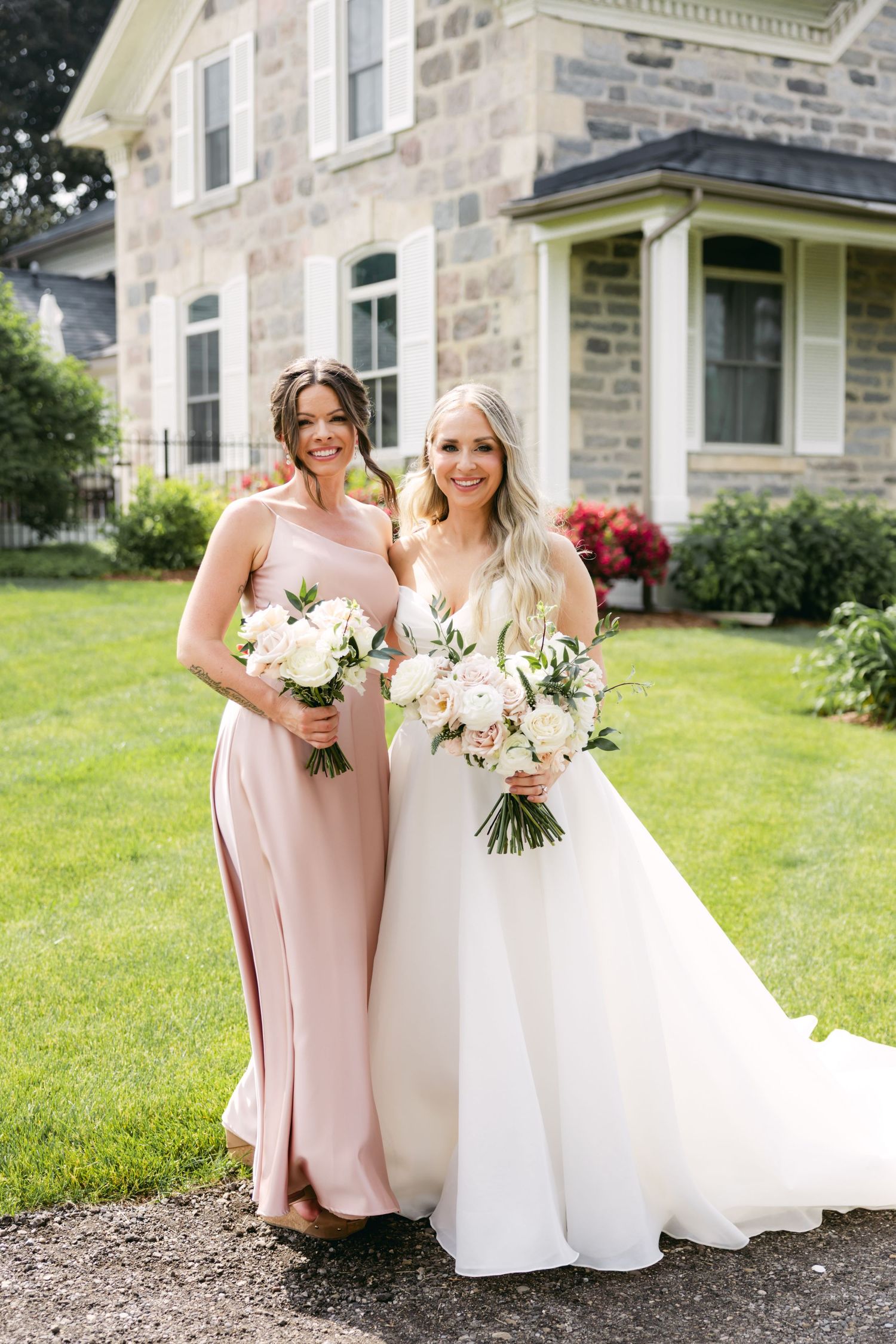 Wedding party members pose with flowers in front of stone building on sunny day.