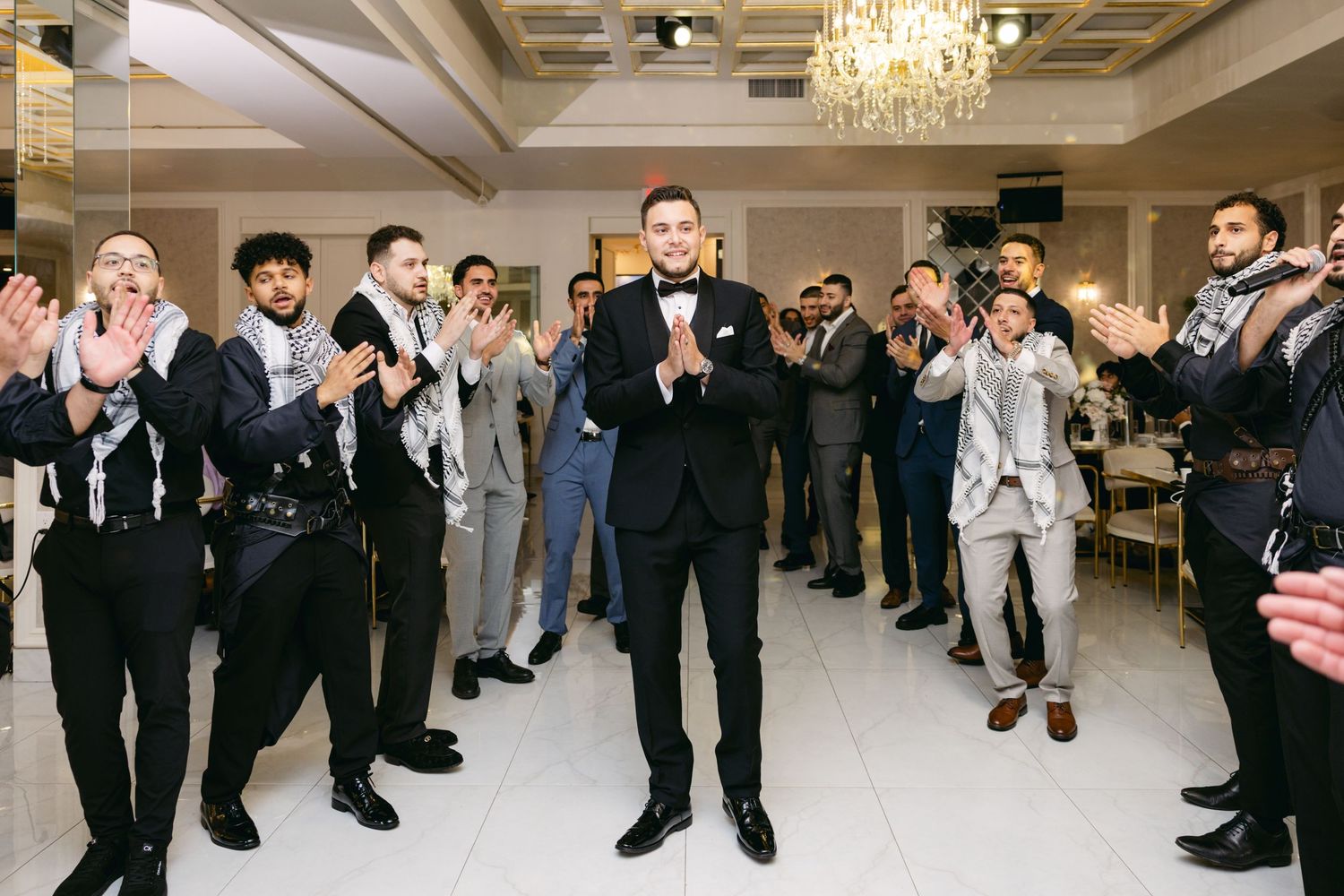 A group of formally dressed guests gather in an elegant ballroom with chandeliers for a celebration.