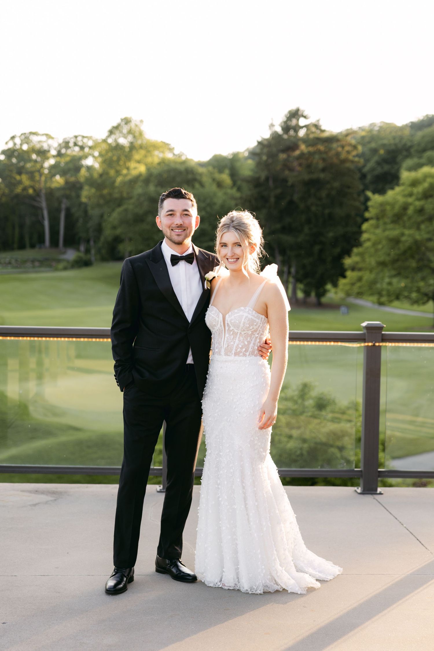 Couple poses on an outdoor terrace overlooking a scenic golf course during golden hour.