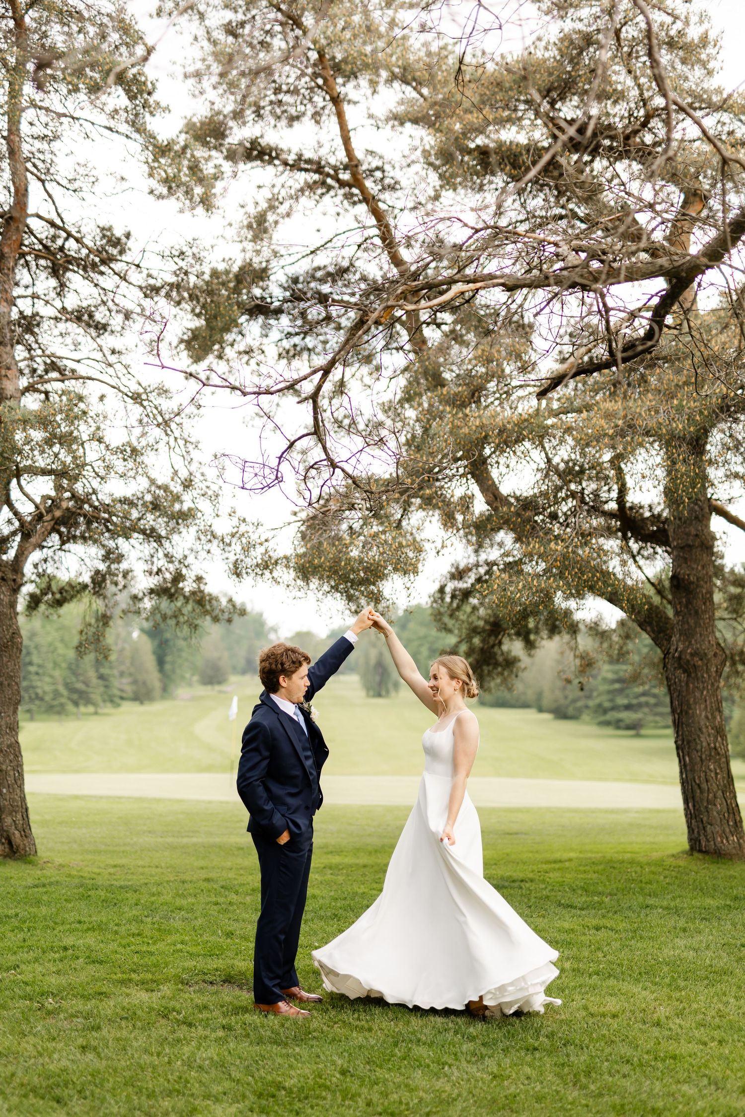 A joyful wedding couple twirls and dances together on a scenic grassy hillside.