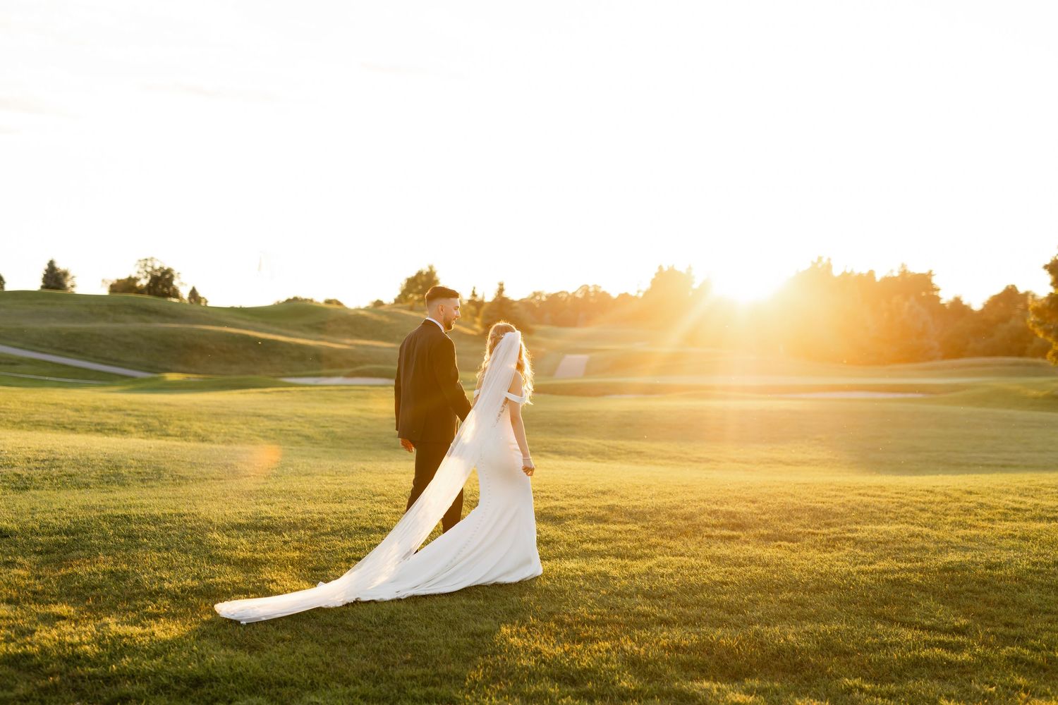 Silhouetted wedding couple walks across a golden-lit hillside at sunset.