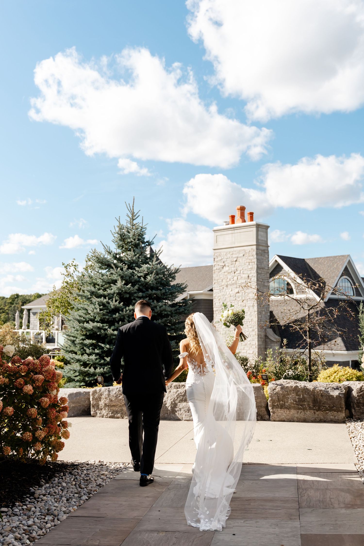 Newlyweds walking on outdoor stone patio with blue sky and elegant architecture.