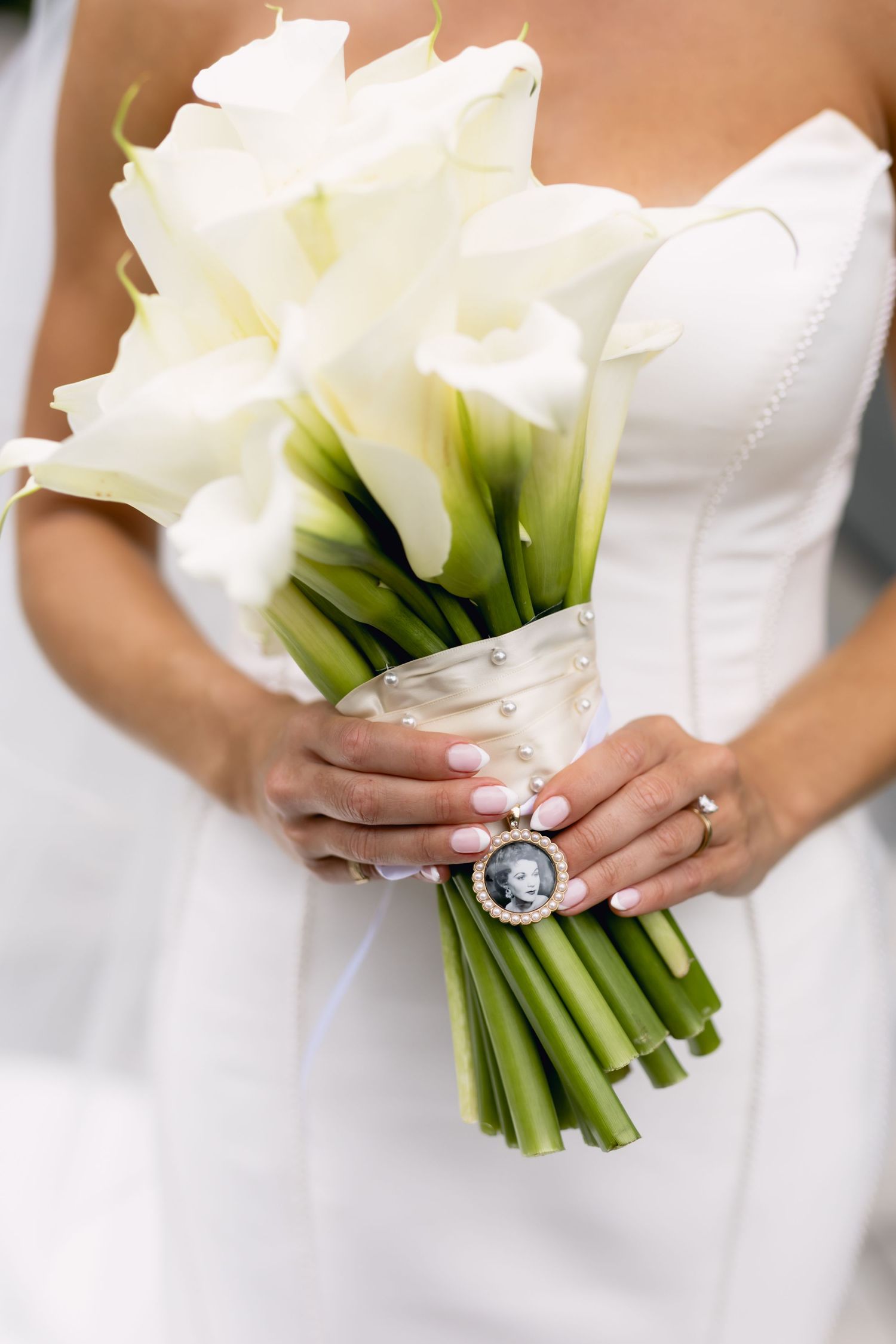 Bride holds elegant white calla lily bouquet with silver memorial photo charm wrapped in ribbon.