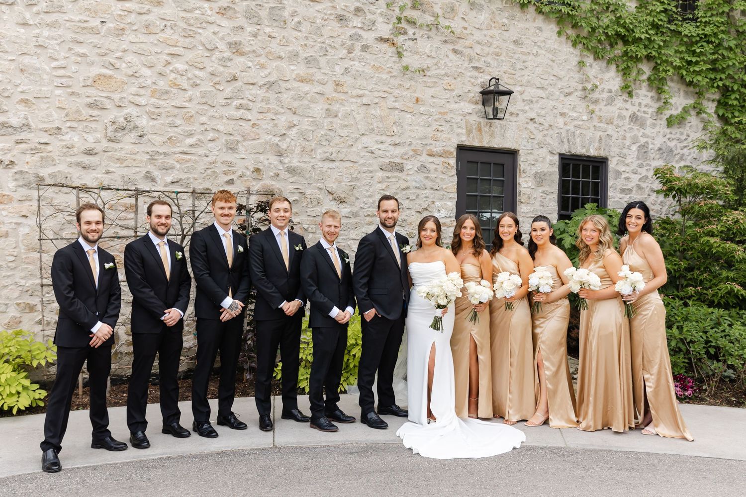 Wedding party poses in front of stone building, bridesmaids in gold gowns and groomsmen in black suits flanking bride and groom.