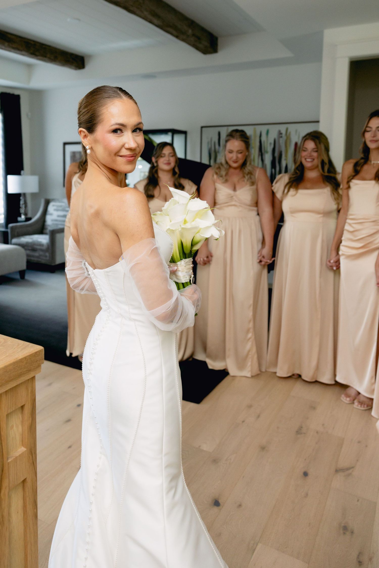 Bride in backless white gown holding calla lily bouquet with bridesmaids in blush dresses behind her.