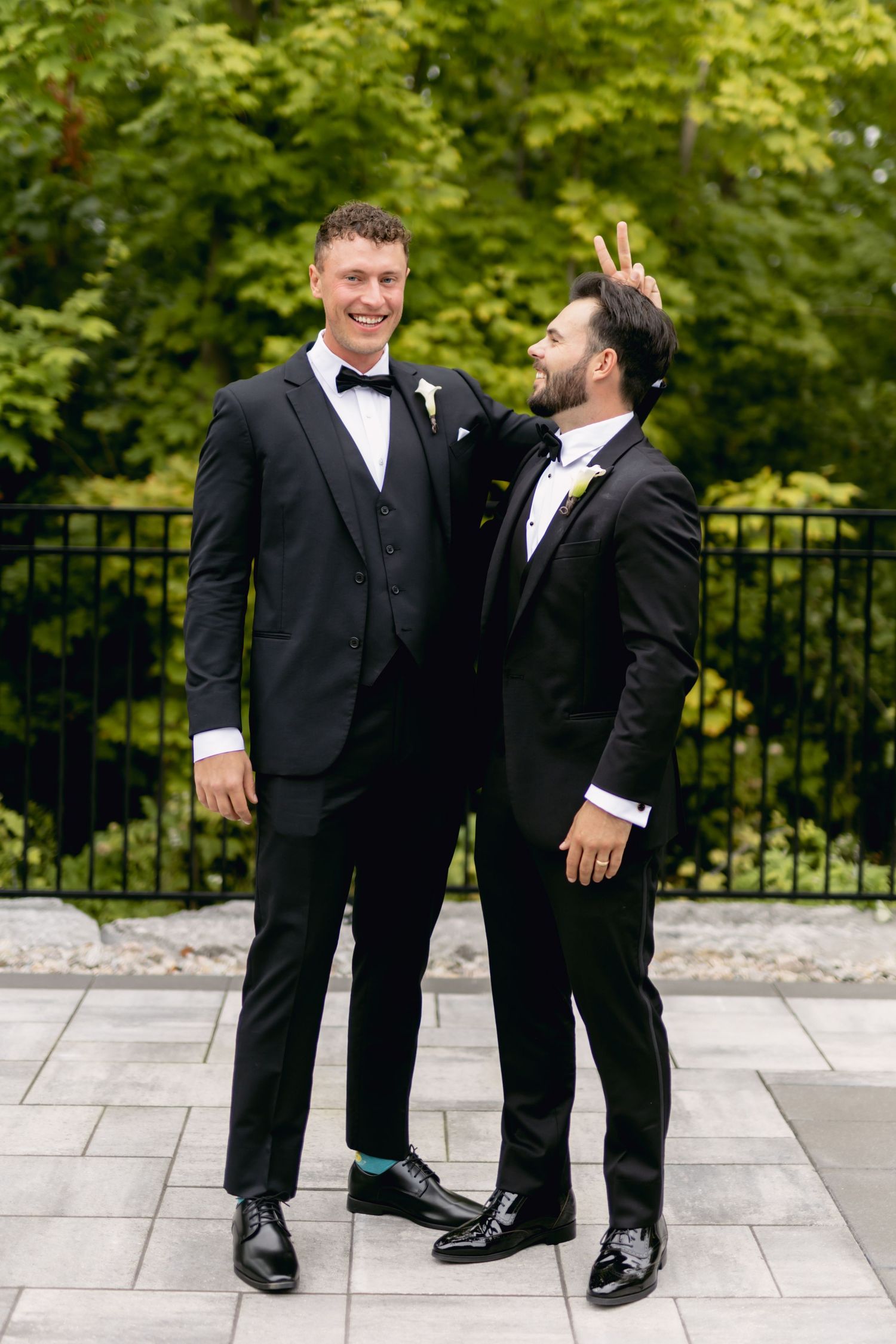 Two groomsmen in black tuxedos pose outdoors, one playfully giving bunny ears to the other.