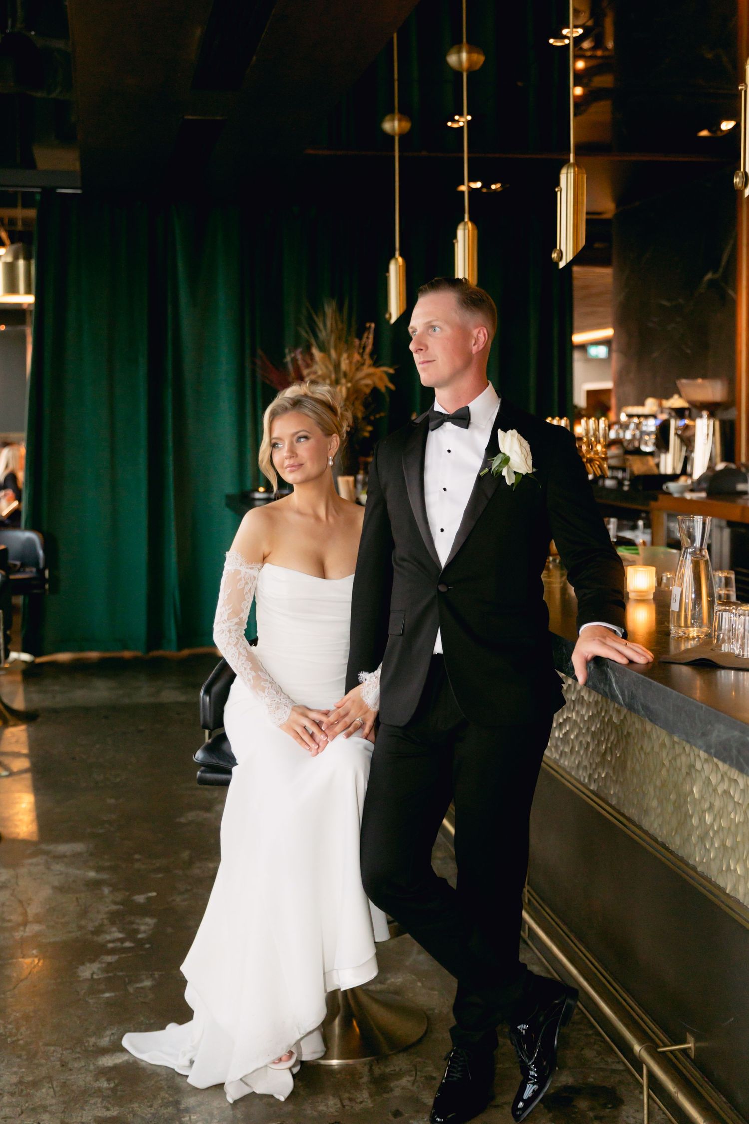 Elegant bride in off-shoulder gown and groom in black tuxedo pose together at a sophisticated dark bar with emerald drapes.
