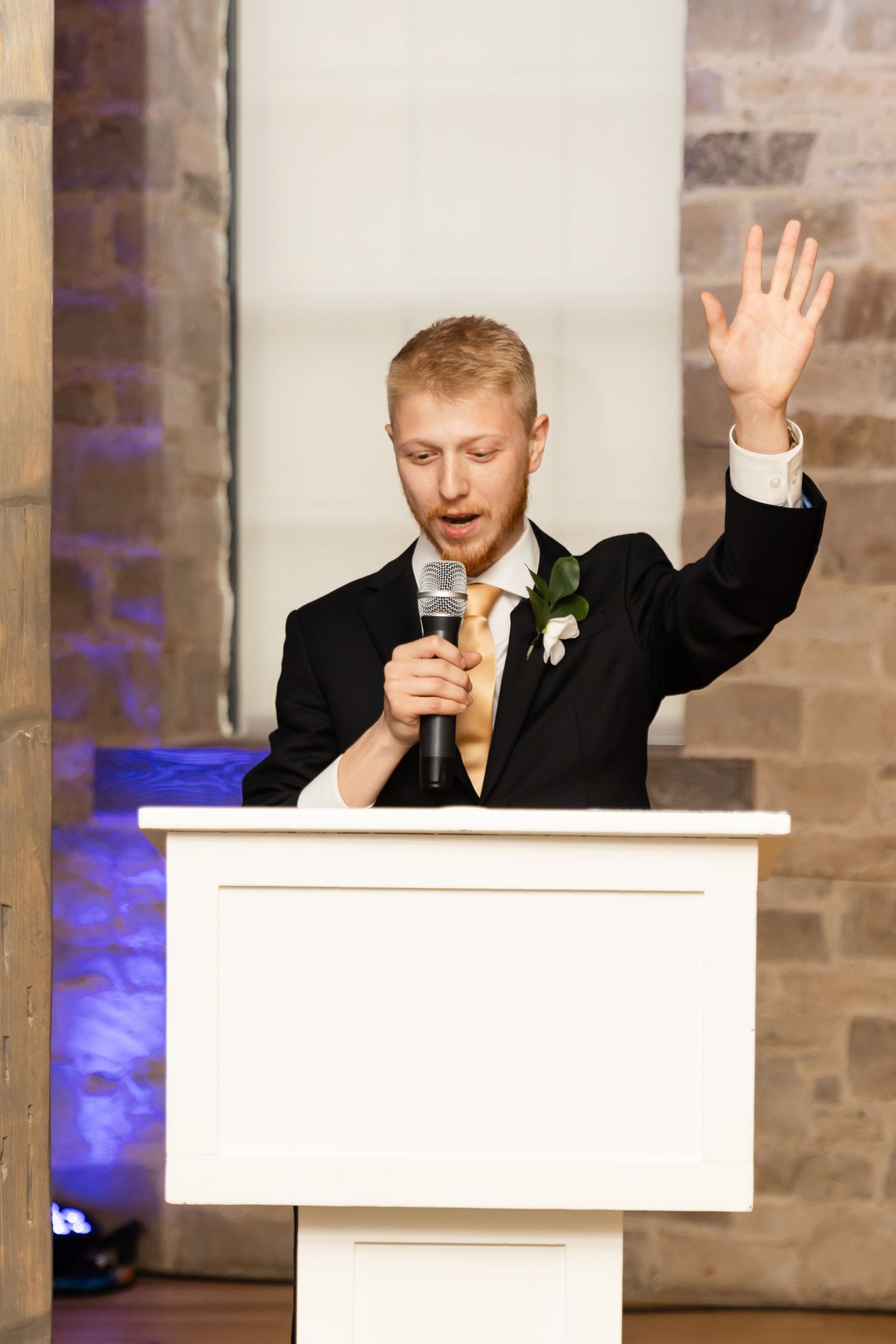 Red-haired groomsman in black suit delivers emotional speech at white podium holding microphone with raised hand.