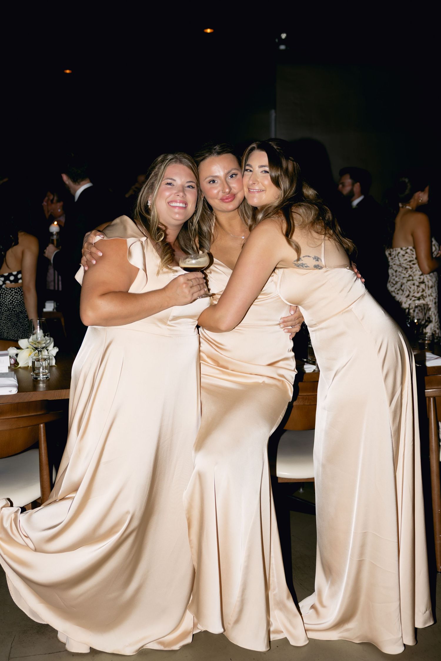 Three bridesmaids in champagne satin gowns hugging and smiling at indoor wedding reception.