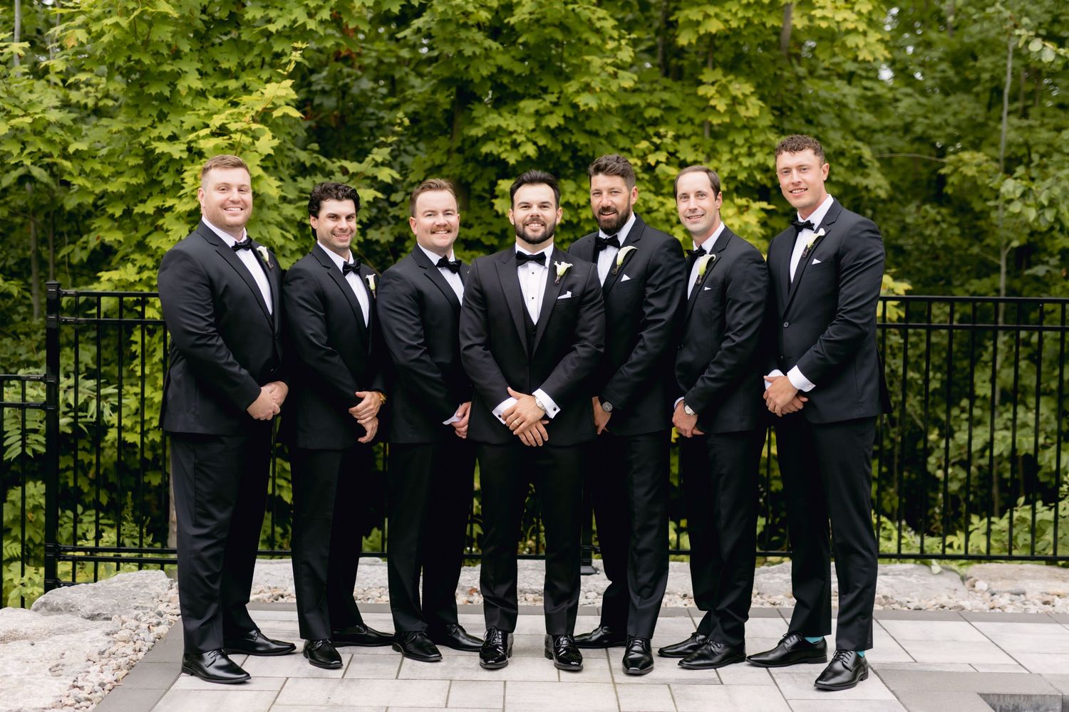 Seven groomsmen in matching black tuxedos posing together outdoors at a wedding celebration.