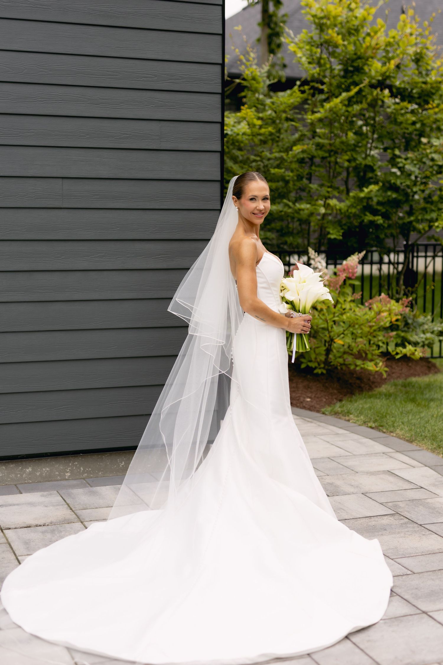 Elegant bride in fitted white gown with long veil holds white calla lily bouquet outdoors.