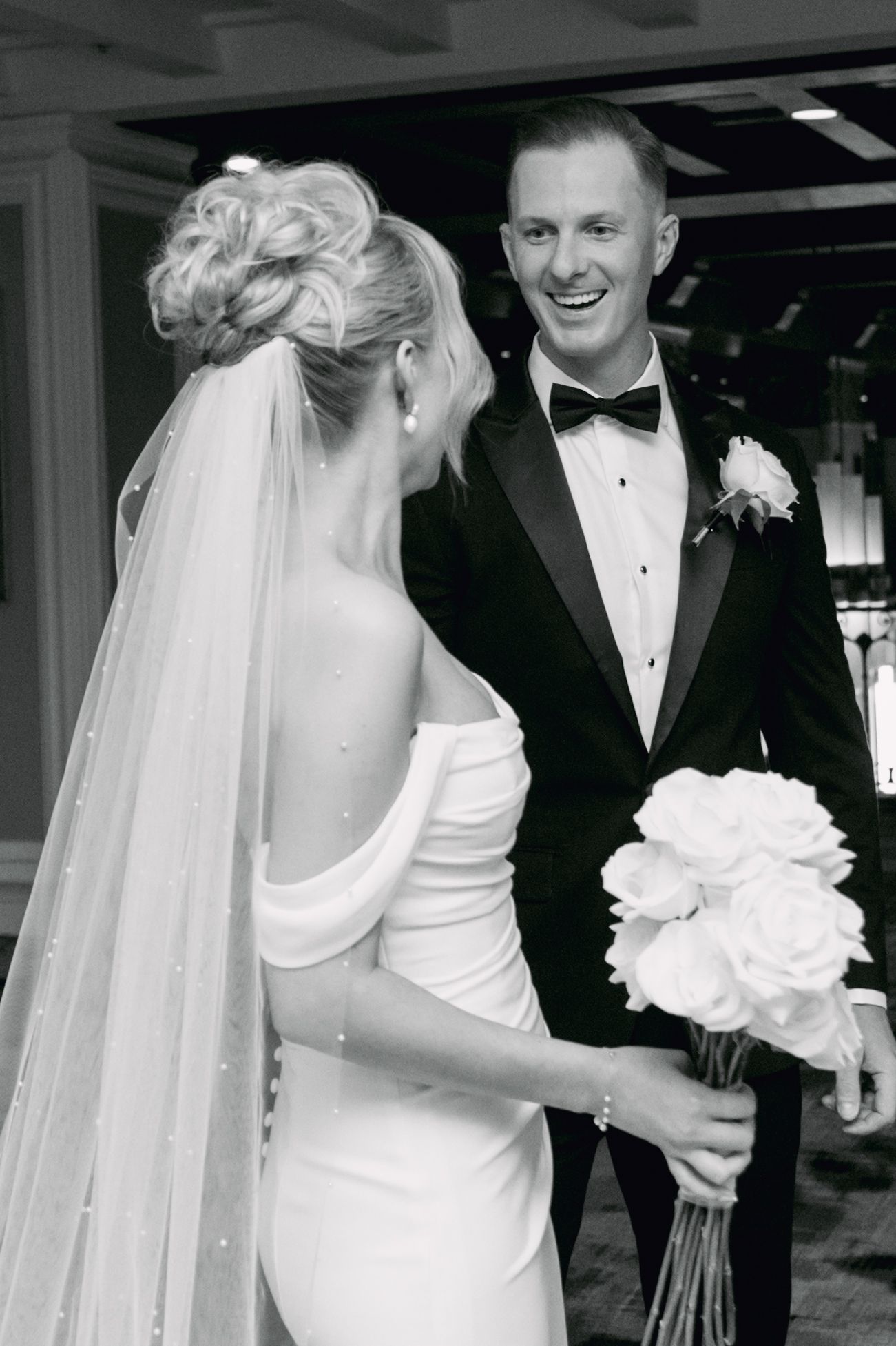 Black and white photo of smiling groom in tuxedo with bride in off-shoulder gown holding white rose bouquet.