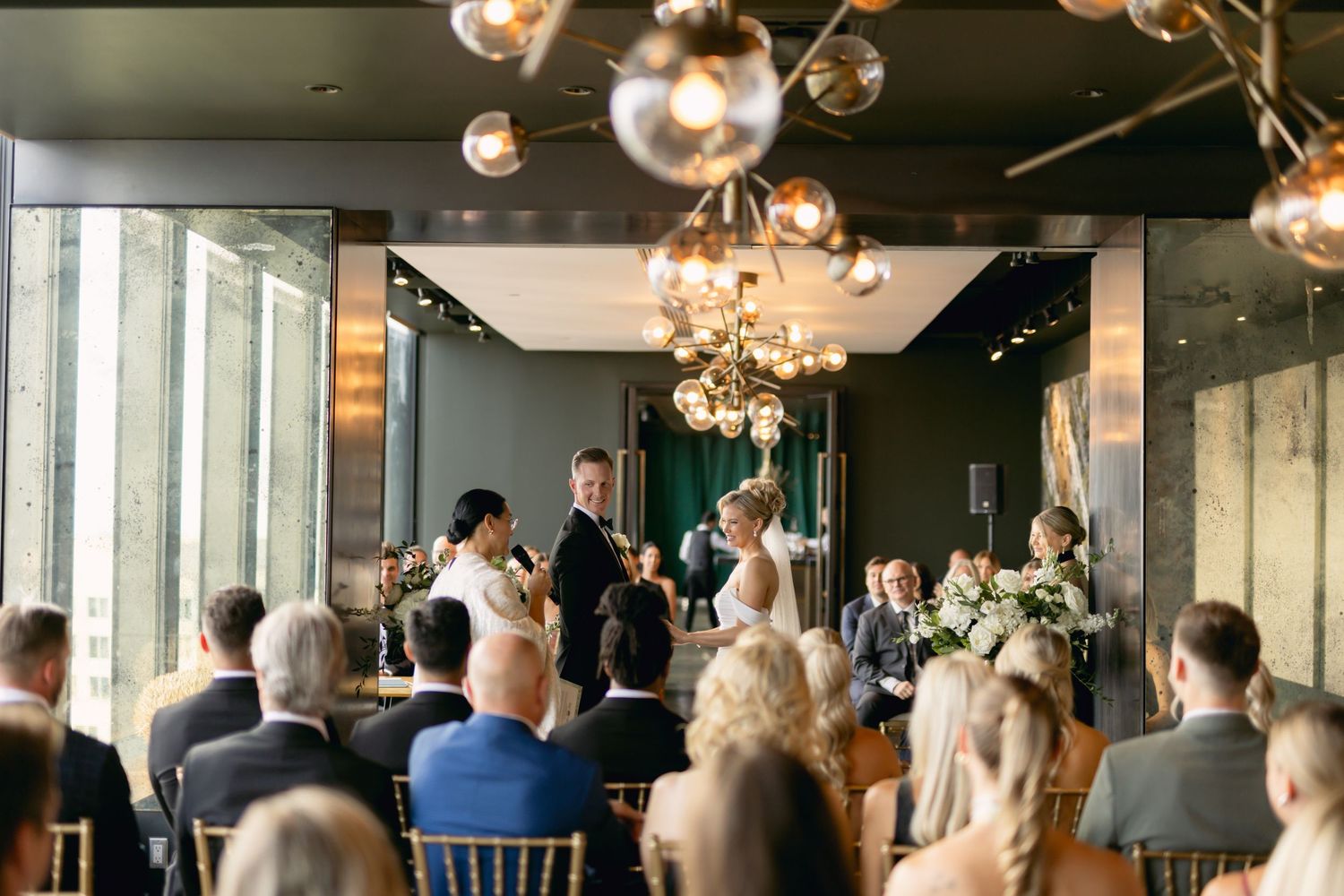 Wedding ceremony in modern venue with gold sputnik chandeliers, guests seated as couple exchanges vows at altar.