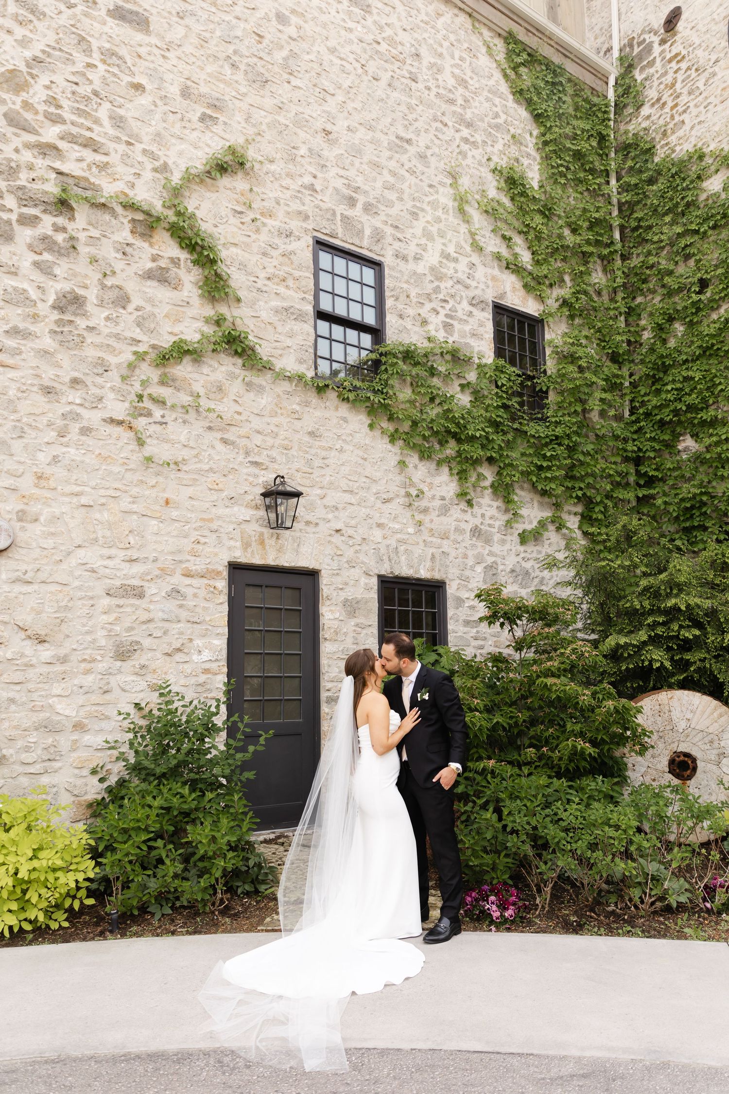 Bride and groom share a romantic kiss in front of a stone building covered in ivy on their wedding day.