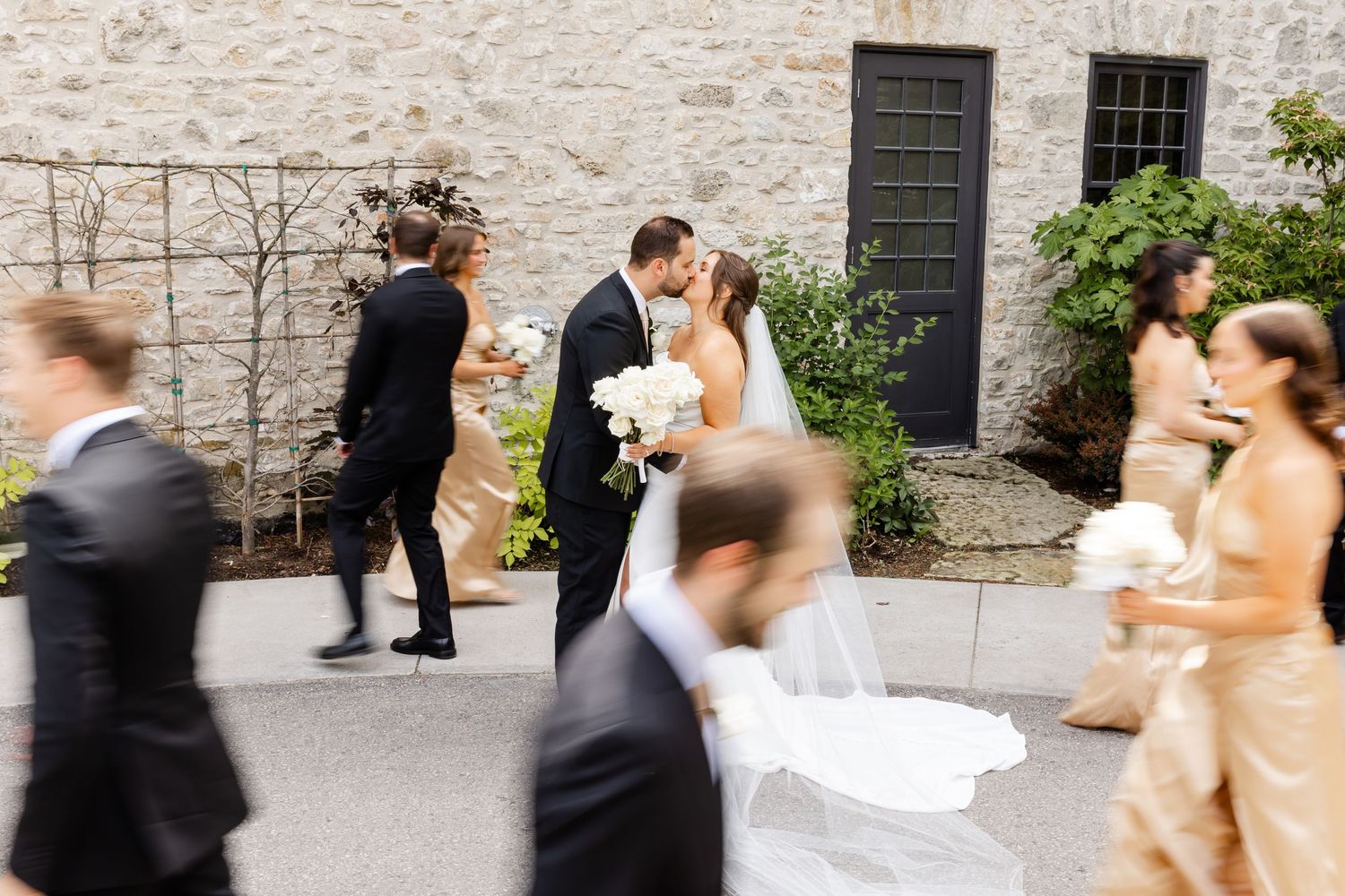 Bride and groom share a kiss surrounded by wedding party in golden gowns and black suits at an elegant outdoor venue.