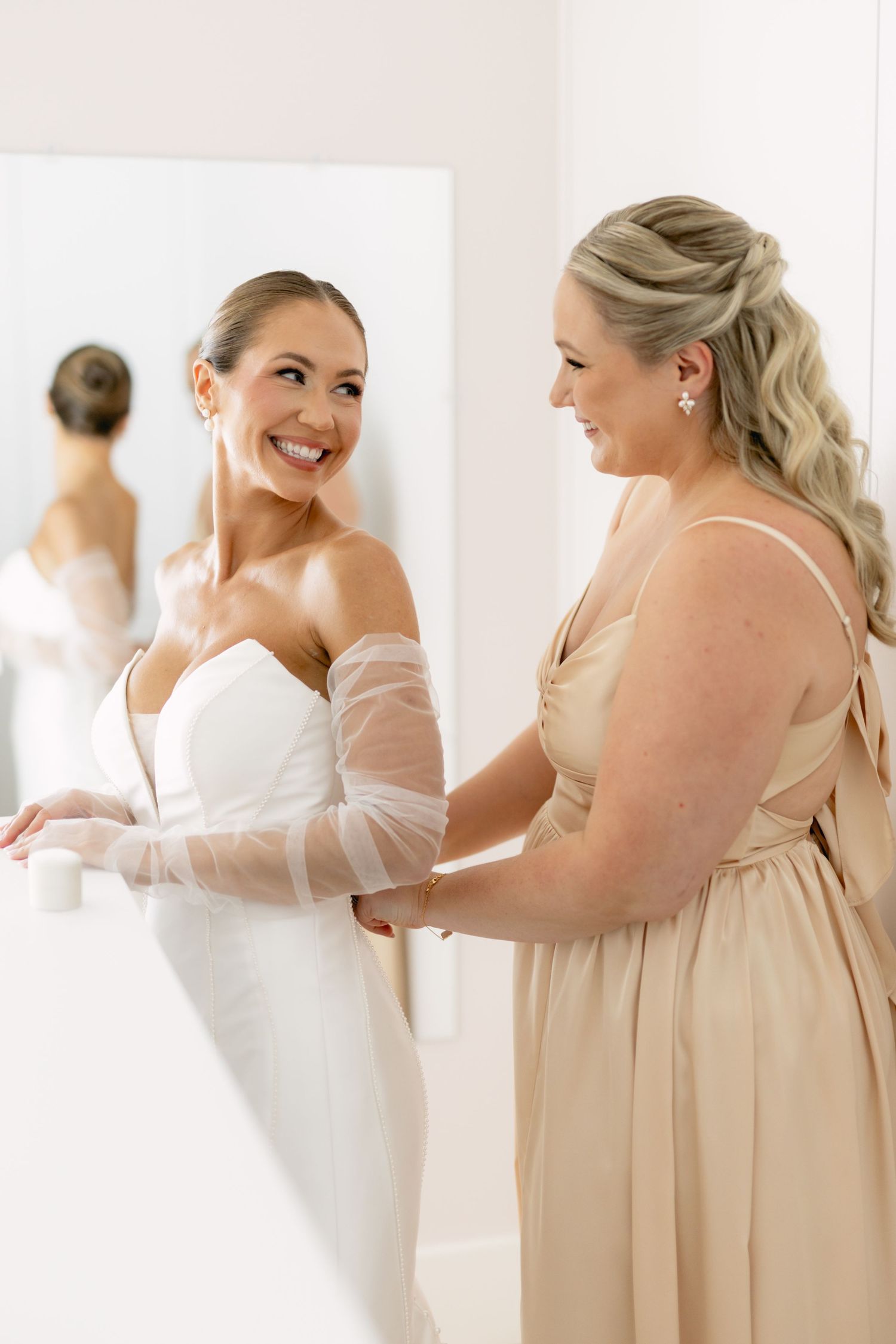 Bride in white gown smiling with bridesmaid in champagne dress during wedding preparations.