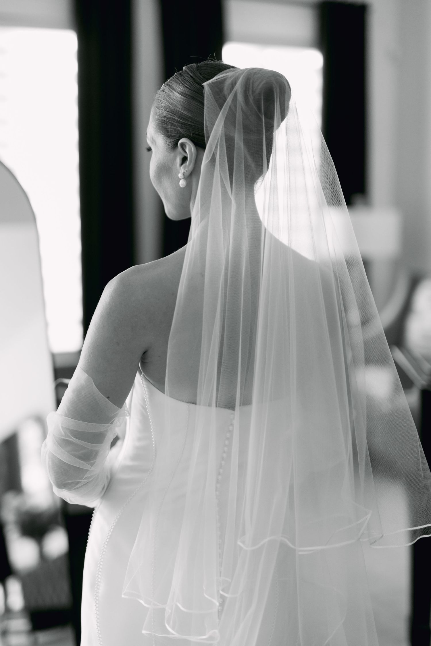 Elegant bride in white gown and veil seen from behind in black and white portrait.