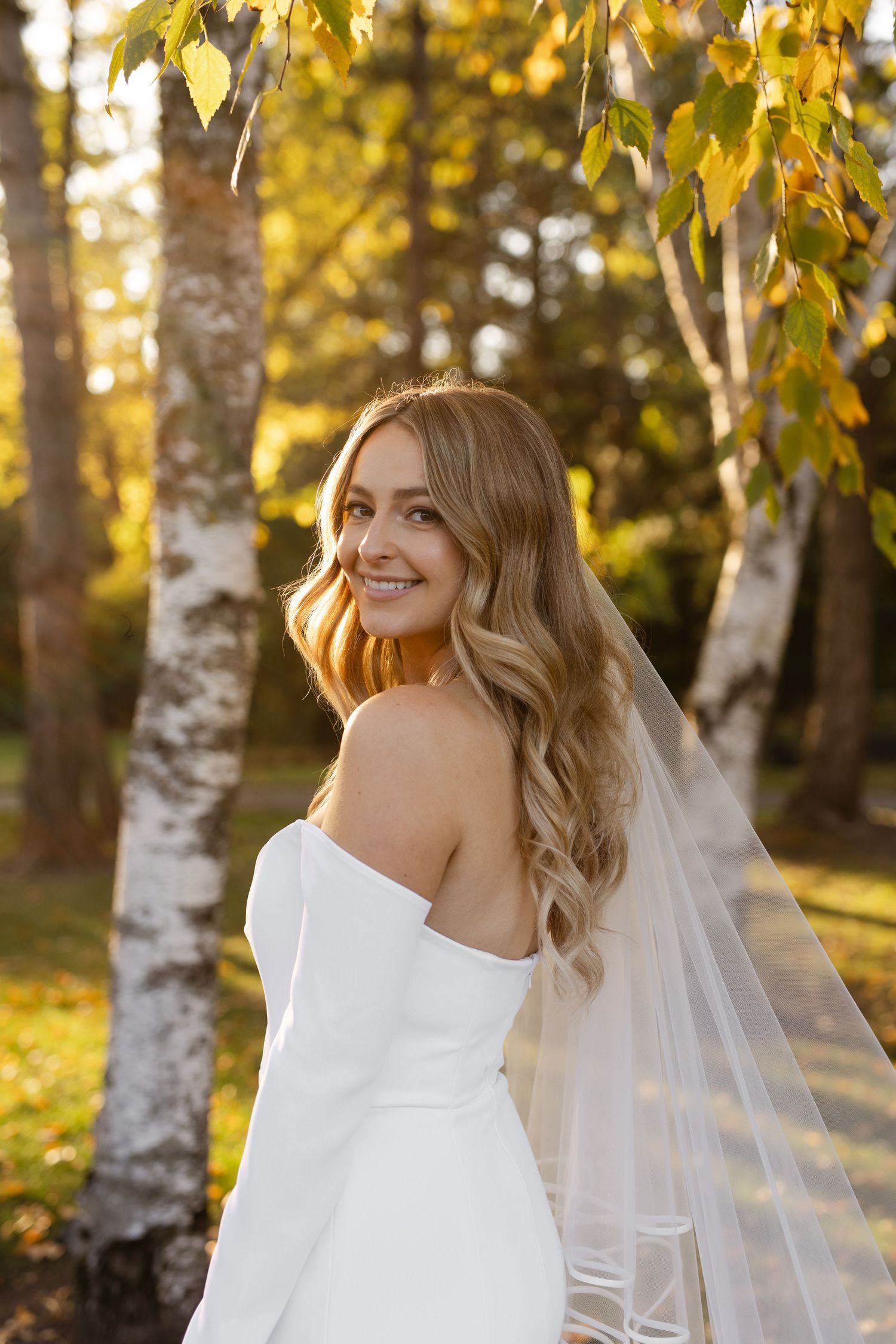 Bride in elegant white dress poses among autumn birch trees in golden light.