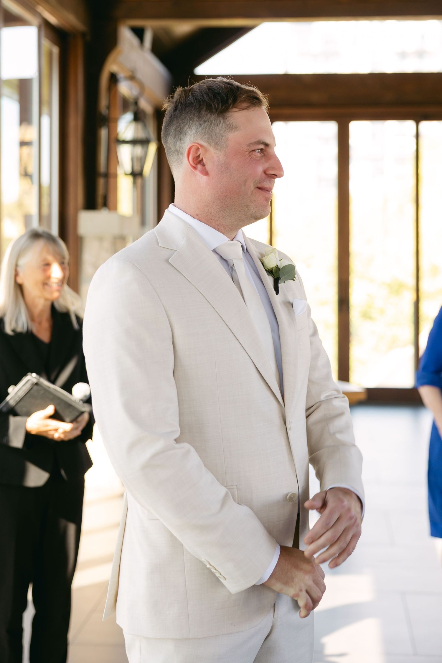 A groom in a cream colored suit stands at attention during a wedding ceremony in an elegant venue.