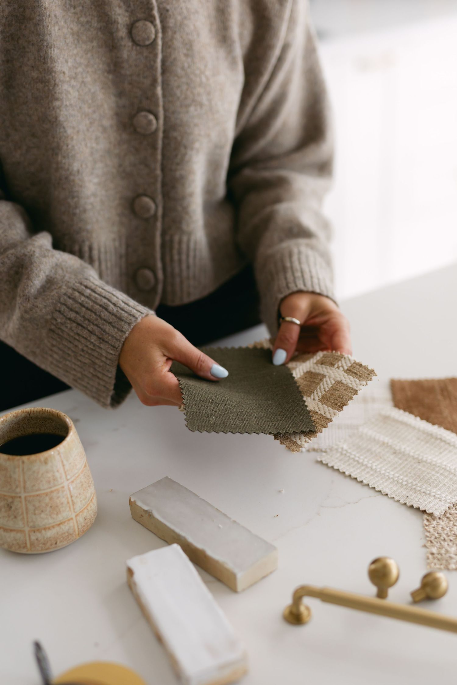 Hands in gray sweater arranging fabric swatches and material samples on white surface.