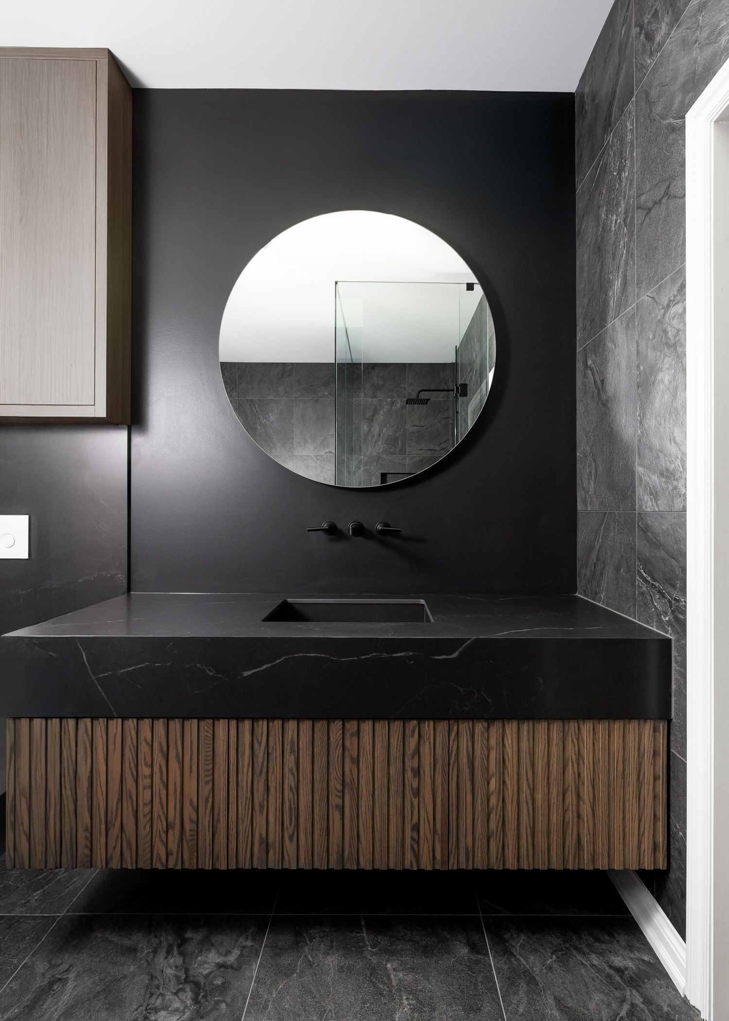 Luxurious black bathroom vanity with wooden slats and circular mirror against dark walls.