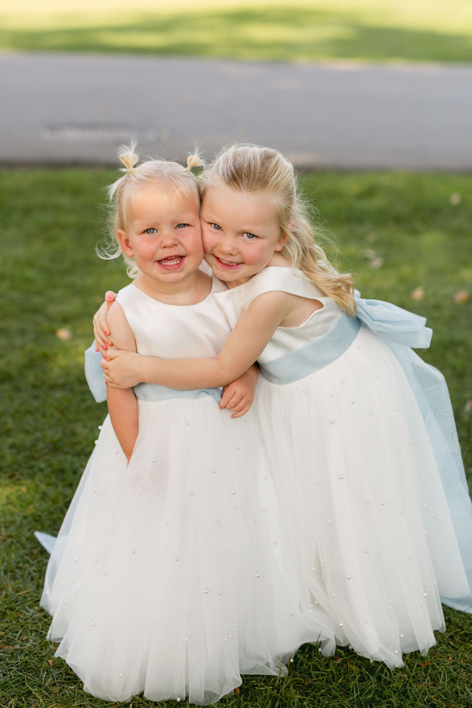 Two flower girls in white tulle dresses hug and smile on a grassy lawn.