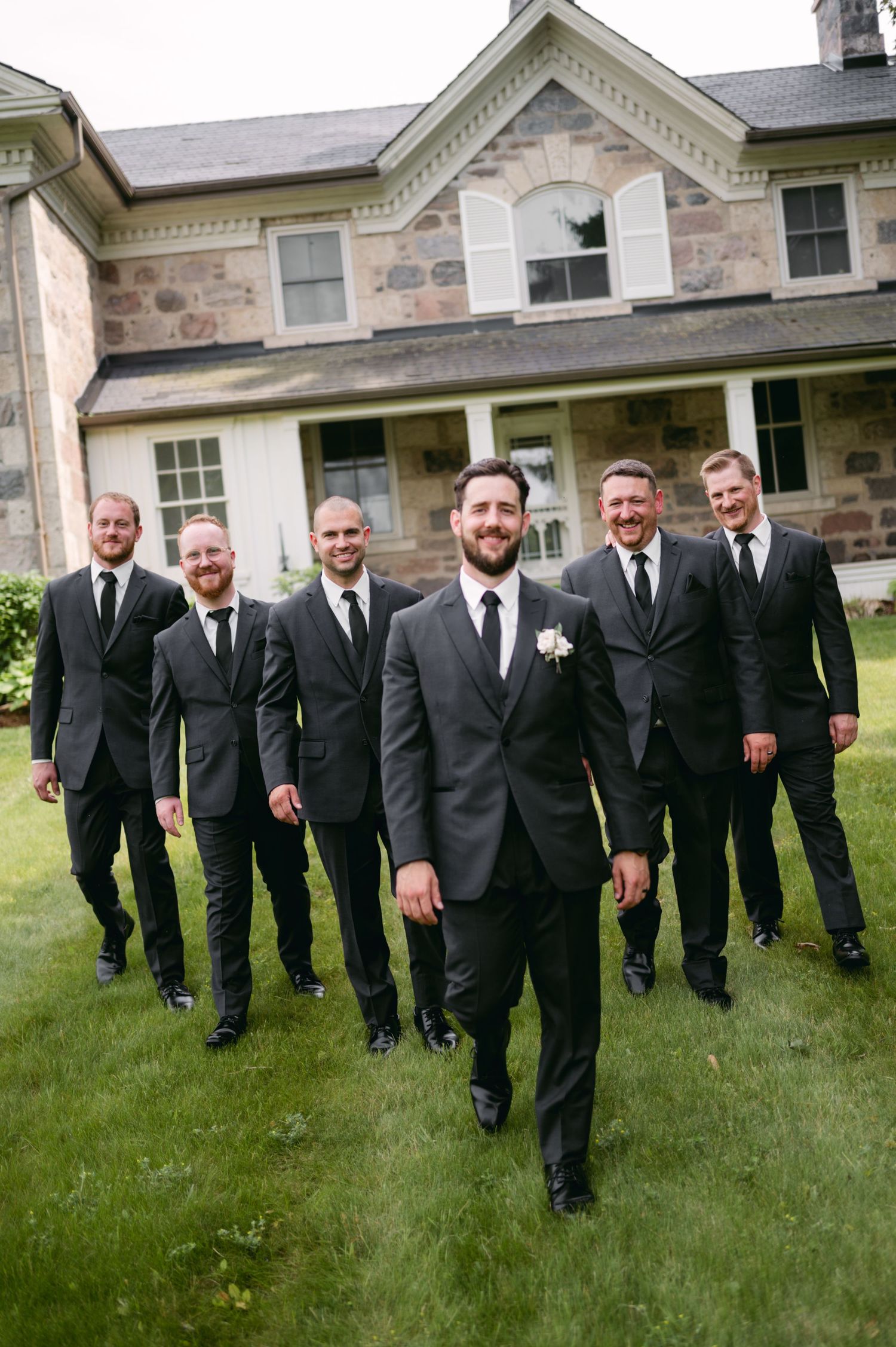 Large group of groomsmen in black suits walk across grass near historic stone building.