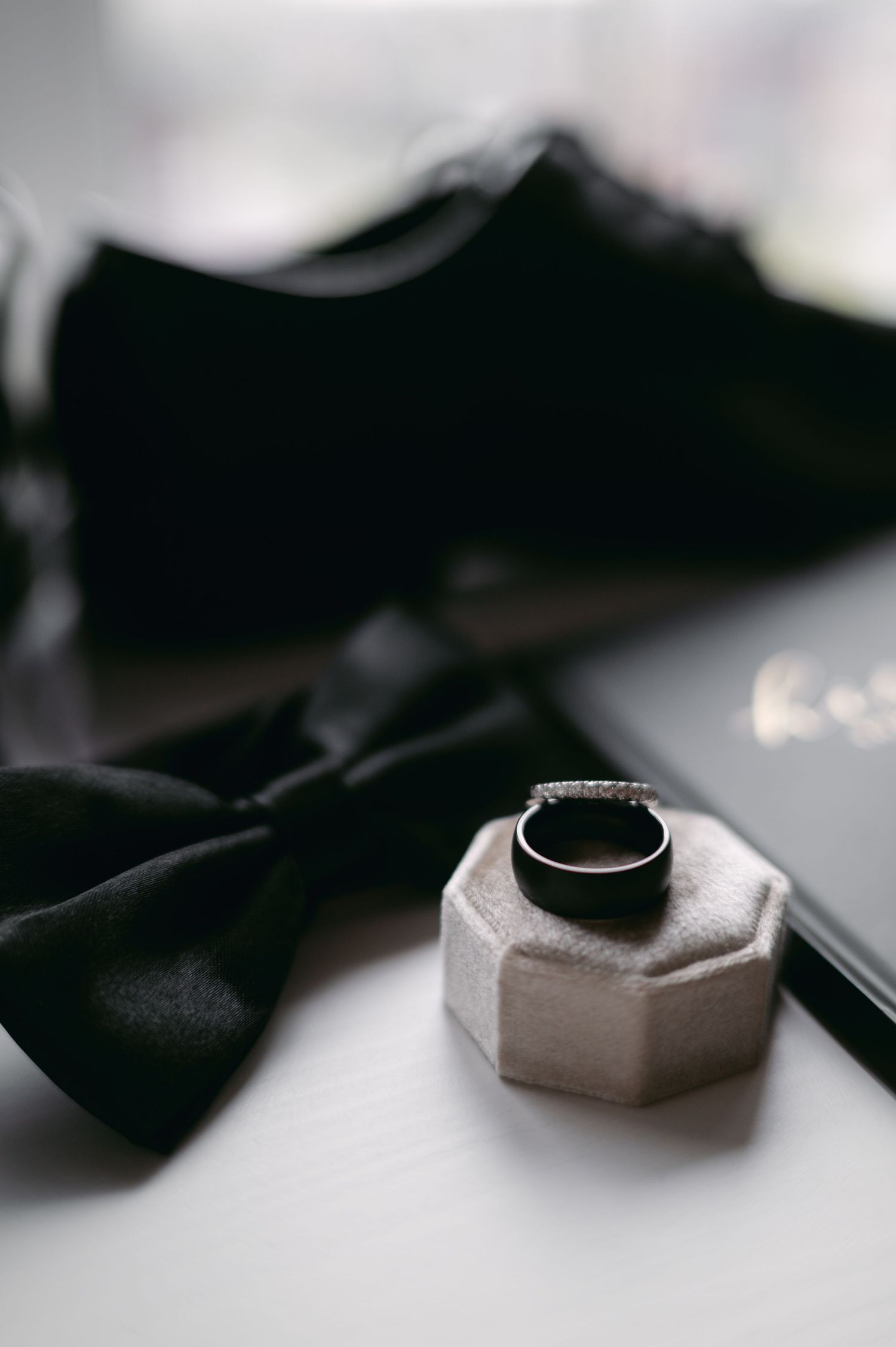 Black bow tie and wedding ring displayed on a hexagonal stand against a light background.