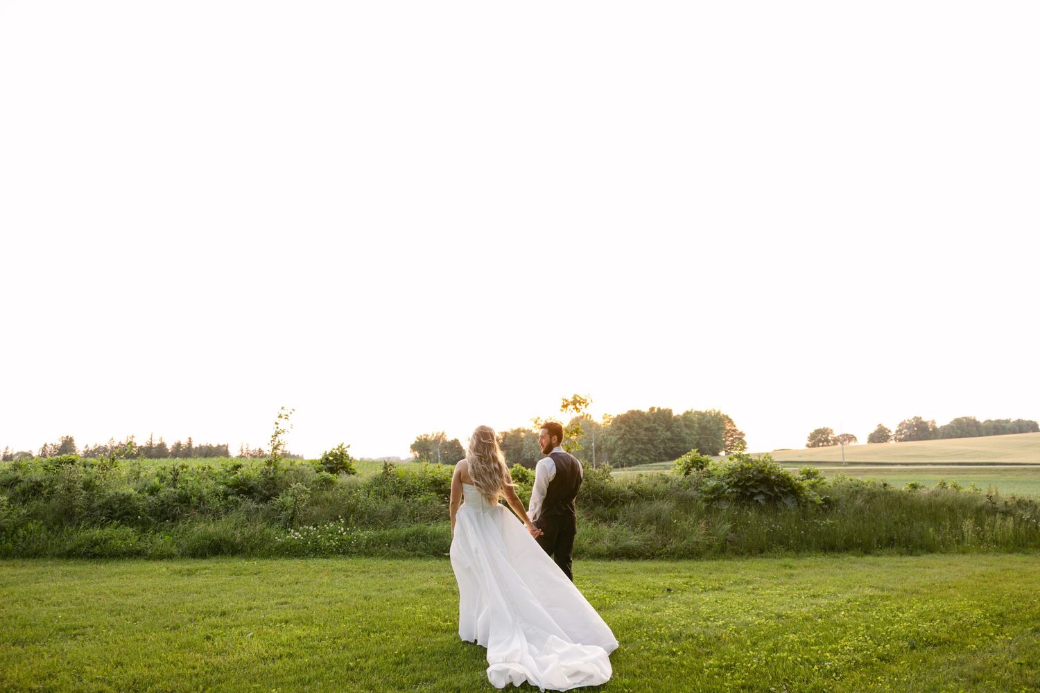 Wedding couple poses playfully in a golden sunset field with white tents in the background.