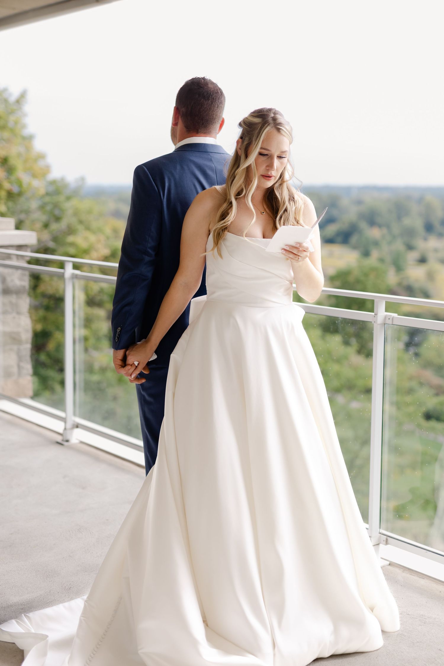 Bride in elegant white gown reads letter during first look moment on balcony.