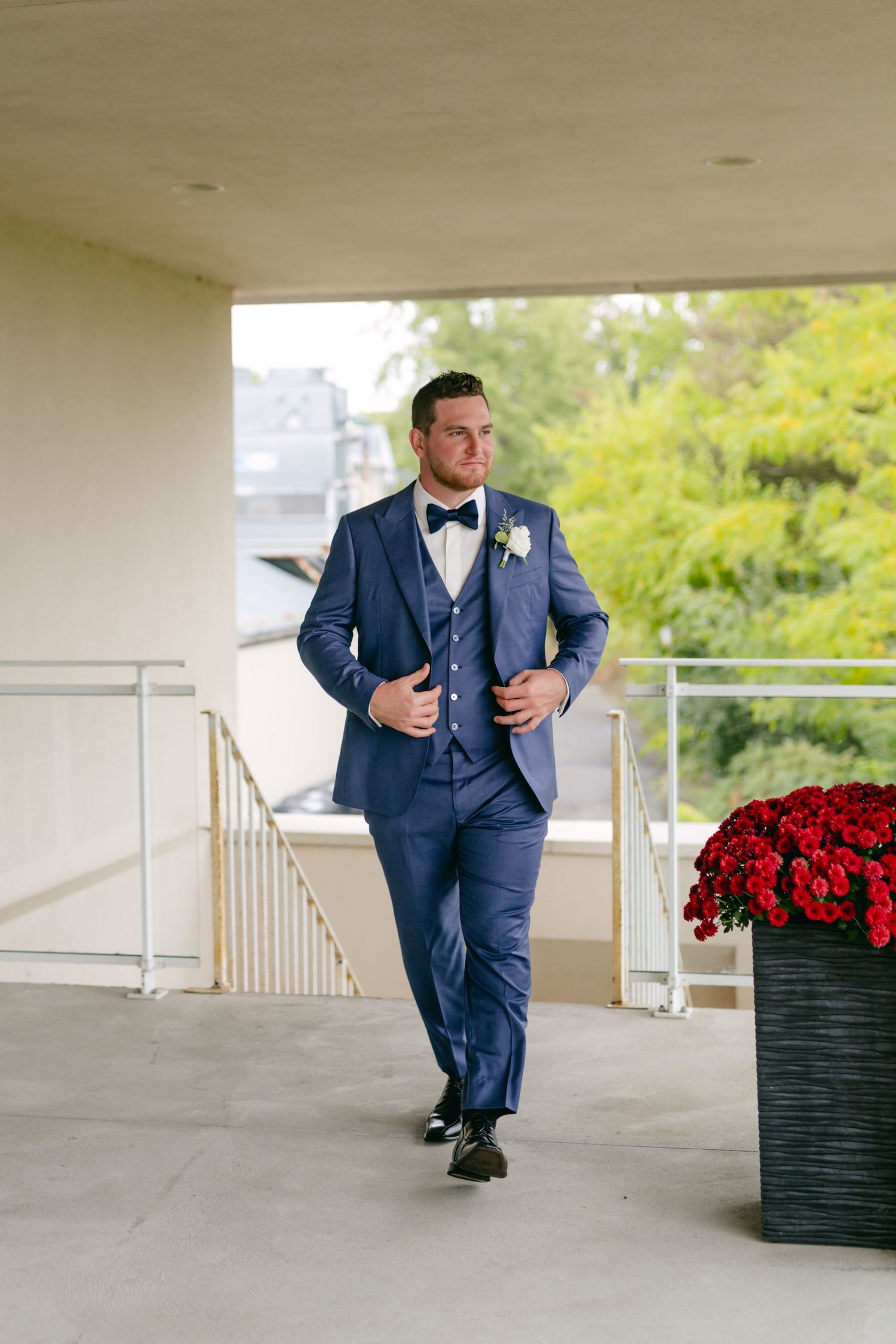 Wedding party member in blue suit walks along covered porch with red floral decorations.