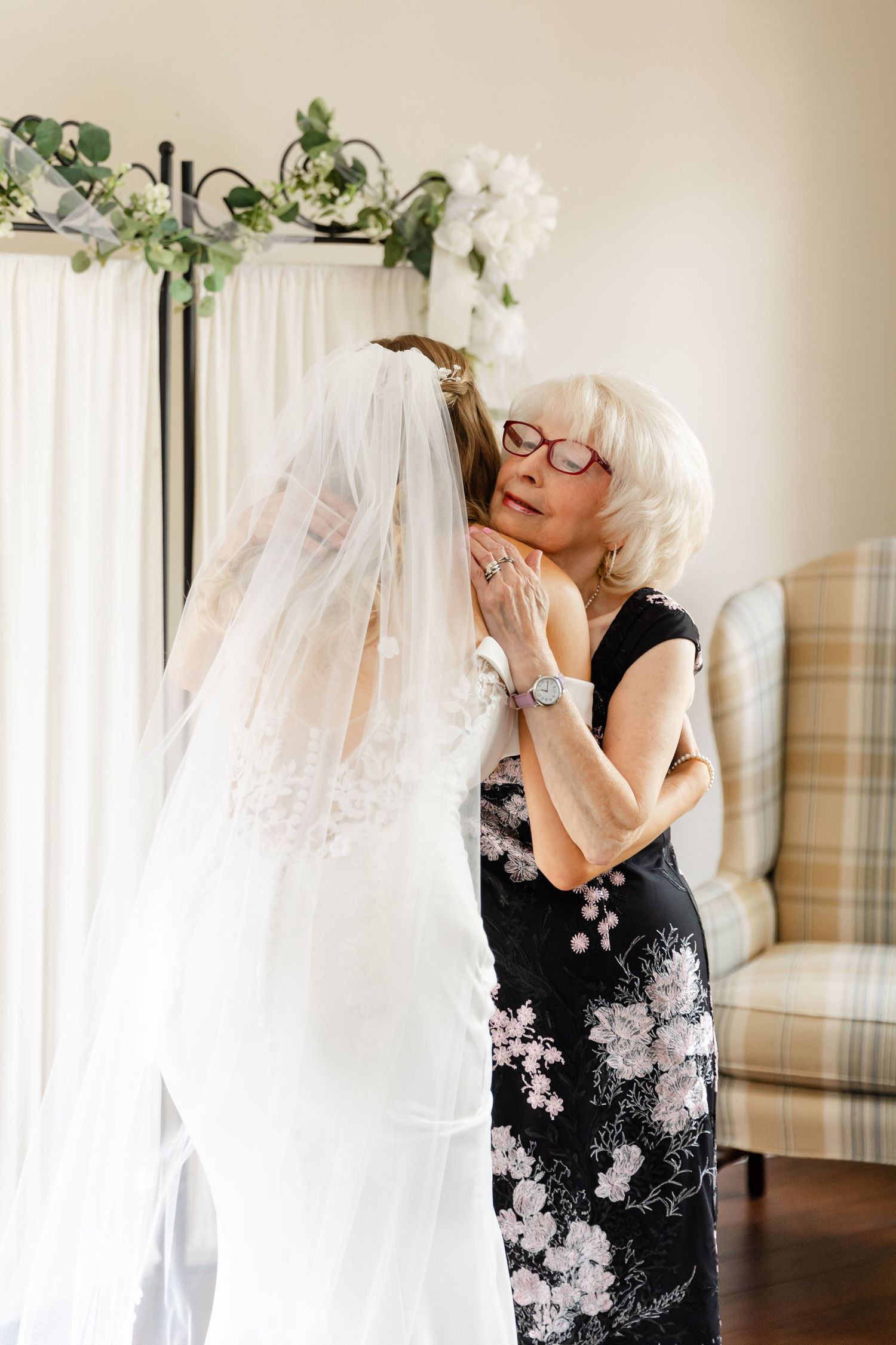 A touching first look moment between a bride and someone in a black floral dress in a sunlit room.