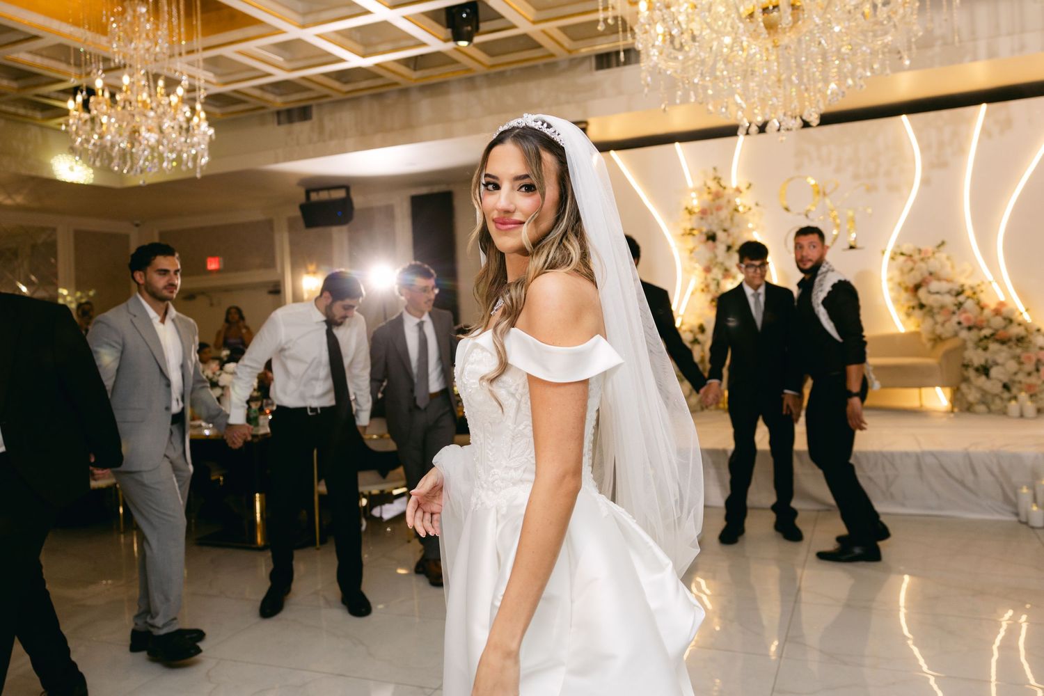 Wedding party dancing and celebrating in an elegant white reception hall with chandeliers.