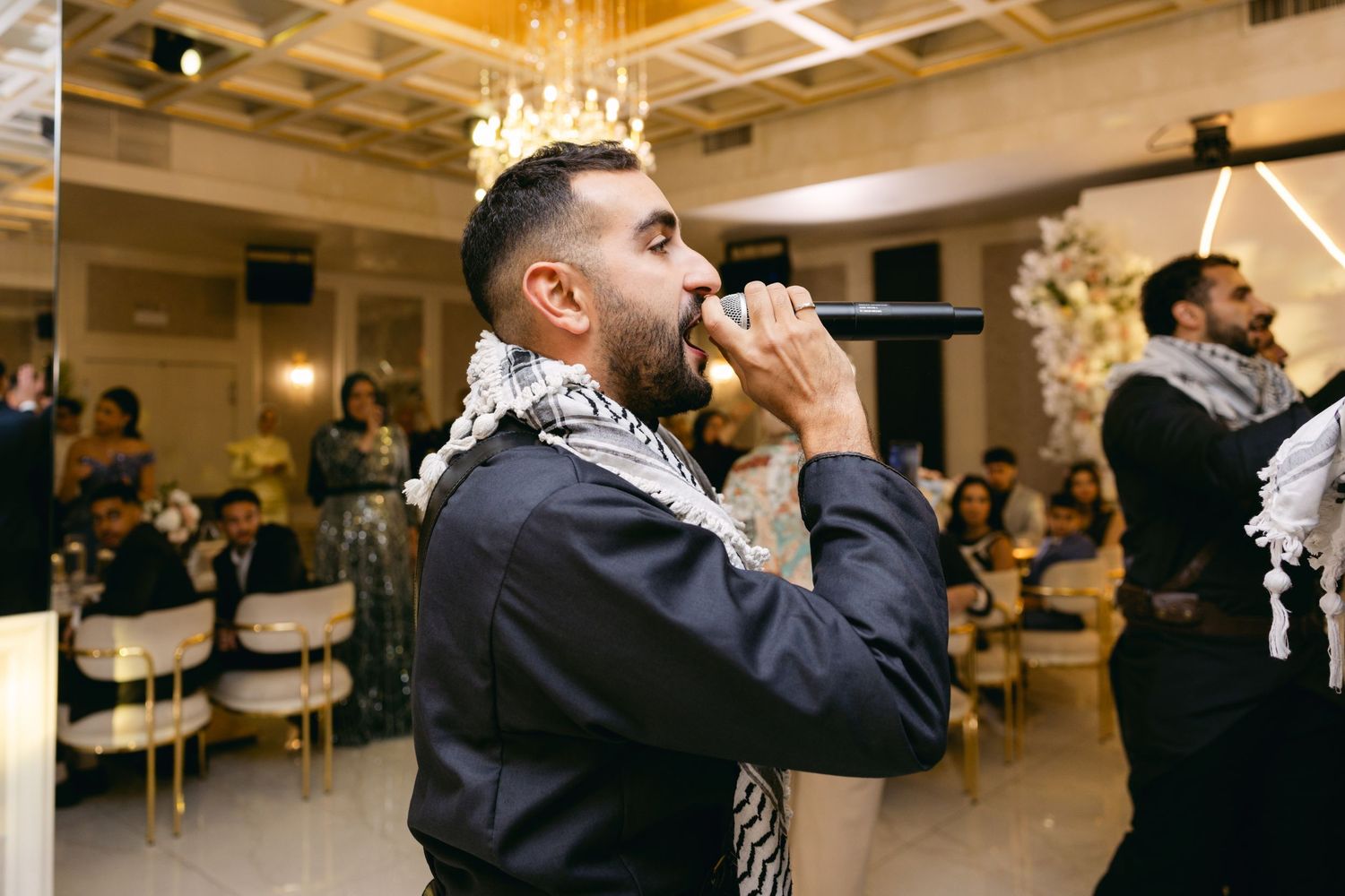 A guest in traditional attire raises a drink during a celebration in an elegant ballroom setting.