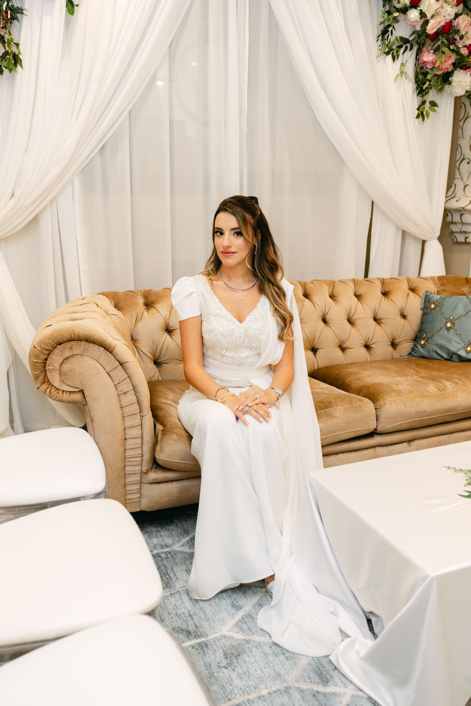 Elegant bride in white wedding jumpsuit sits on vintage tufted beige sofa with flowing cape and floral backdrop.