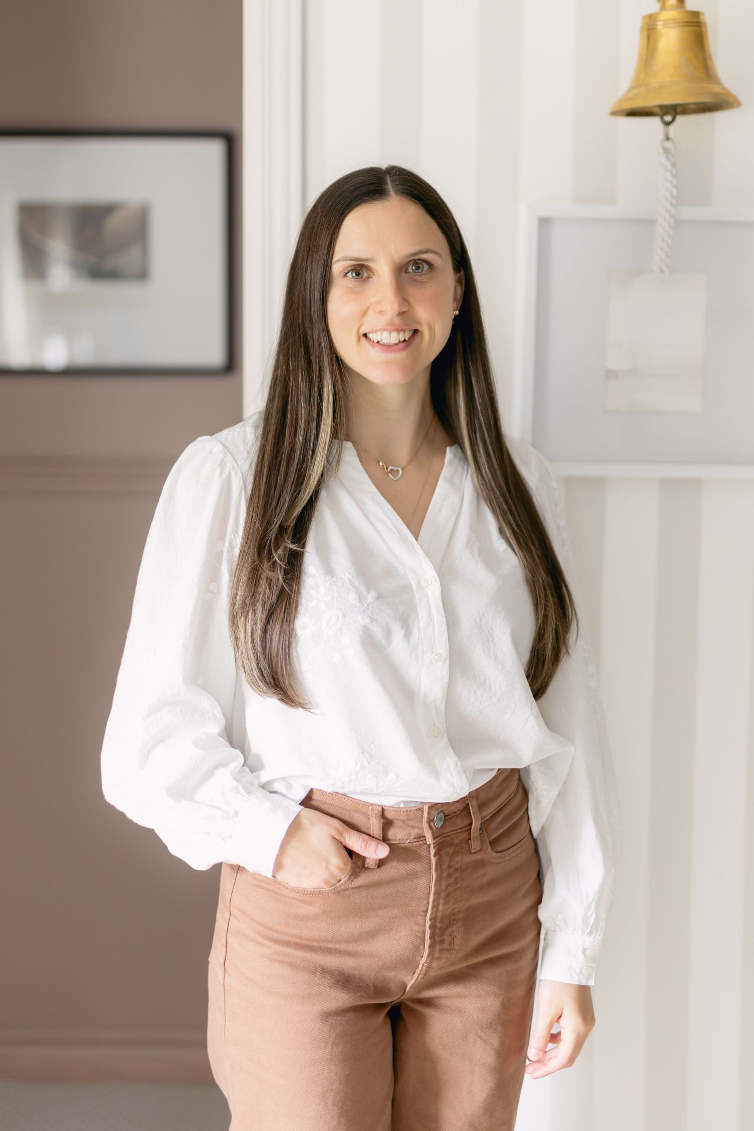 Woman in white blouse and tan pants poses against light colored wall in professional photoshoot series.