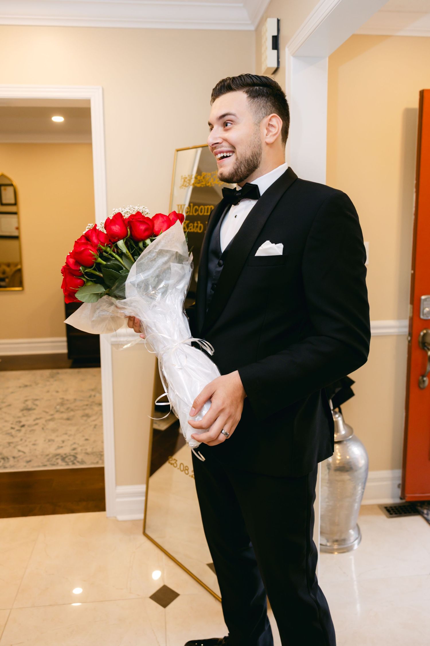 Groom in black tuxedo holding red and white floral bouquet in bright indoor setting.