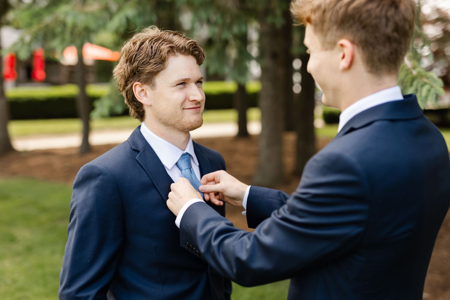 Groomsmen help adjust each other's boutonnieres and ties outdoors before the ceremony.