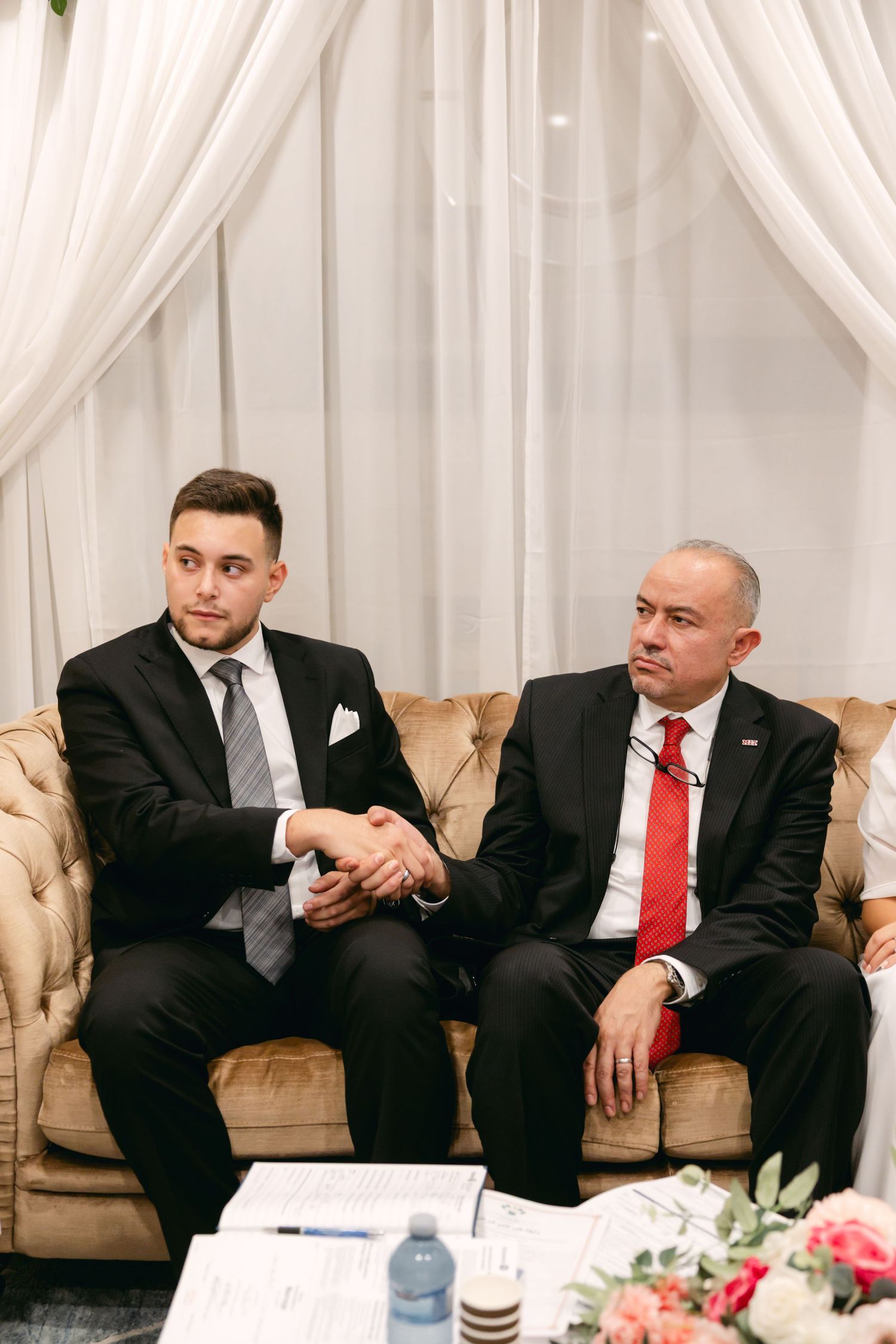 Two people in suits shake hands while seated on a tan leather couch during a formal ceremony.