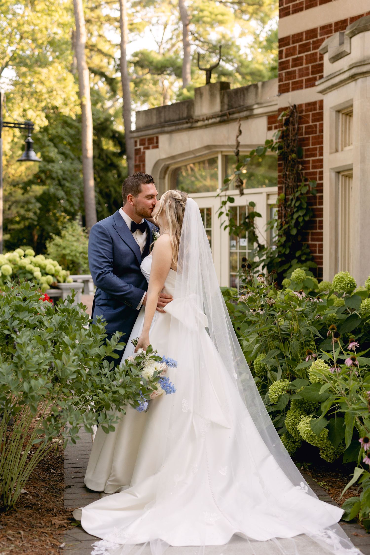 Bride and groom pose together along garden path beside classic brick architecture.
