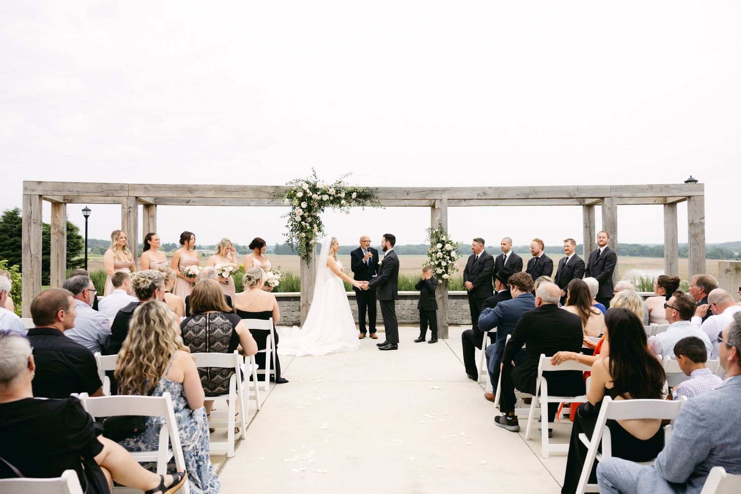 Wedding ceremony guests seated outdoors under a wooden pergola decorated with white floral arrangements.