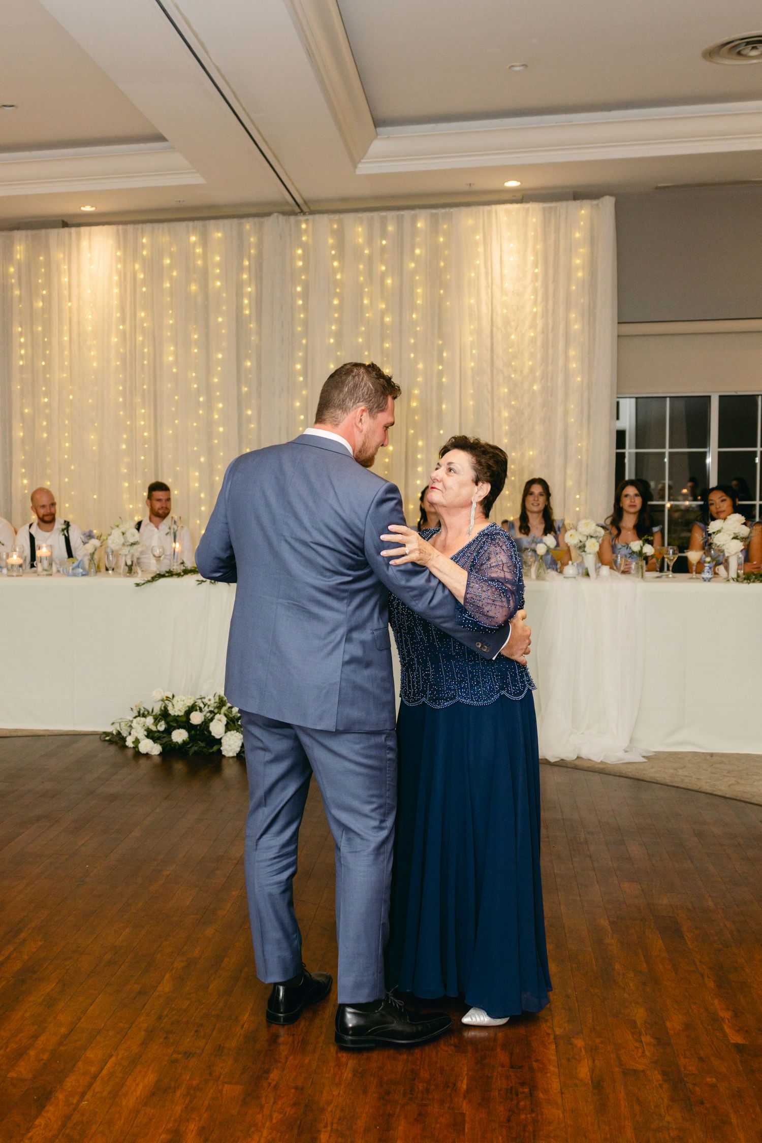 Couple sharing an intimate dance moment on a wooden floor with twinkling fairy lights in background.